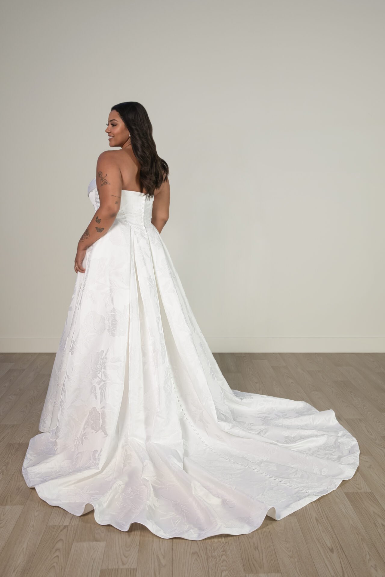 Woman with tattoos in a strapless white wedding gown, facing sideways in a room with light-colored walls and wooden floor.
