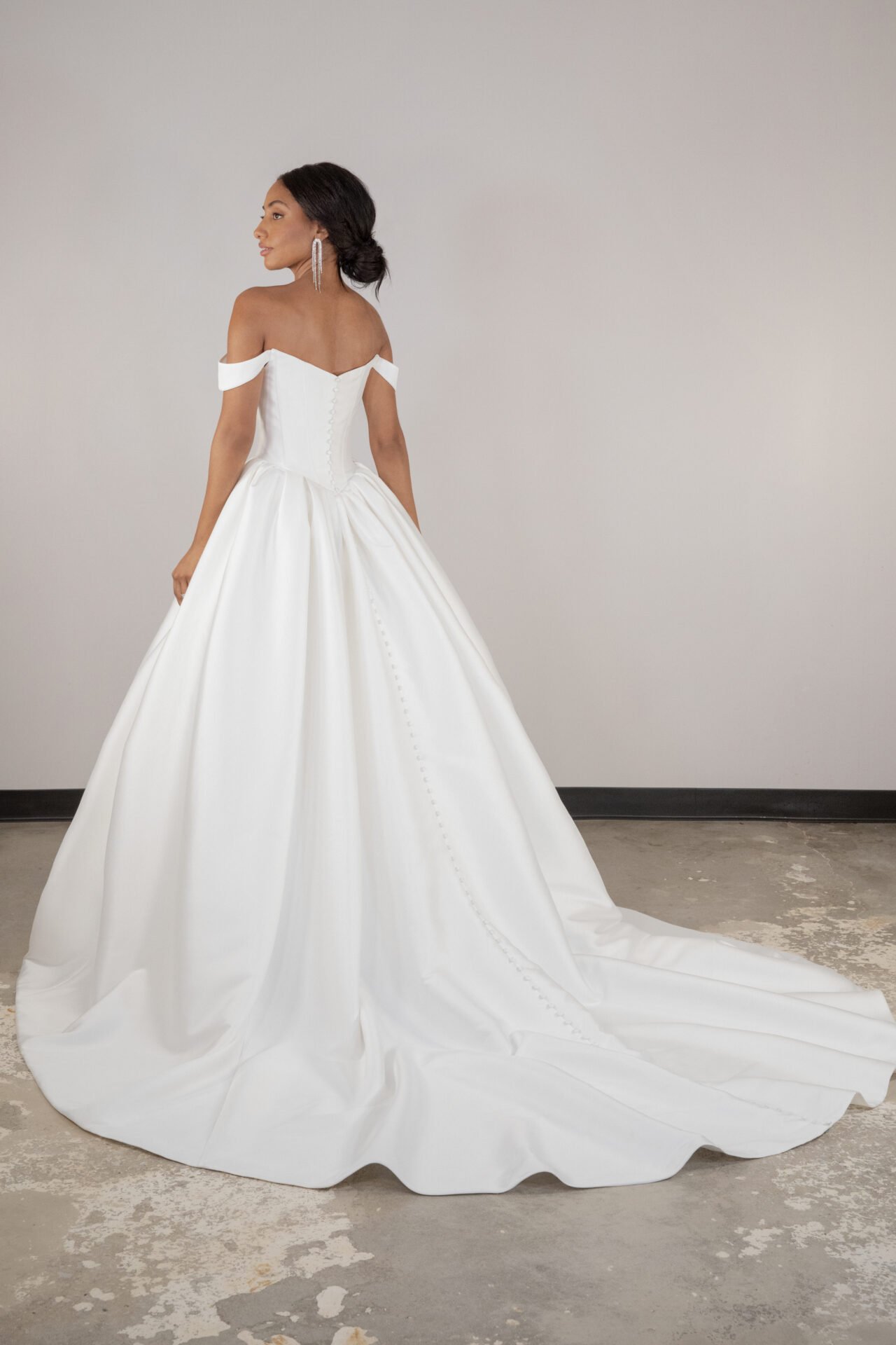 A woman in a white off-the-shoulder wedding gown with a full skirt and train, standing in front of a plain wall.