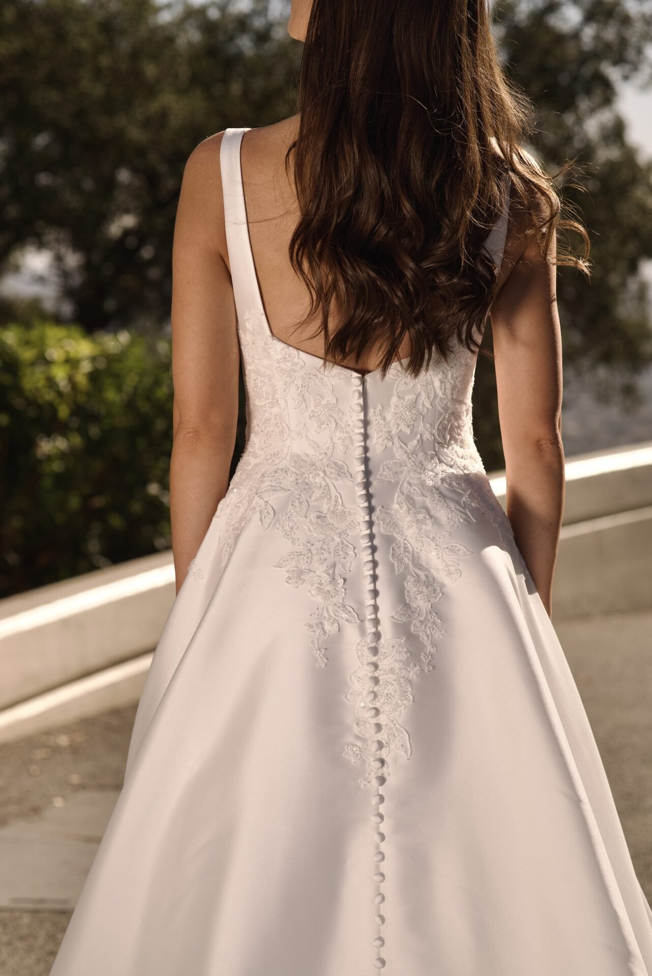 Back view of a woman wearing a white wedding dress with lace details and fabric-covered buttons running down the back, outdoors with trees and a white railing in the background.