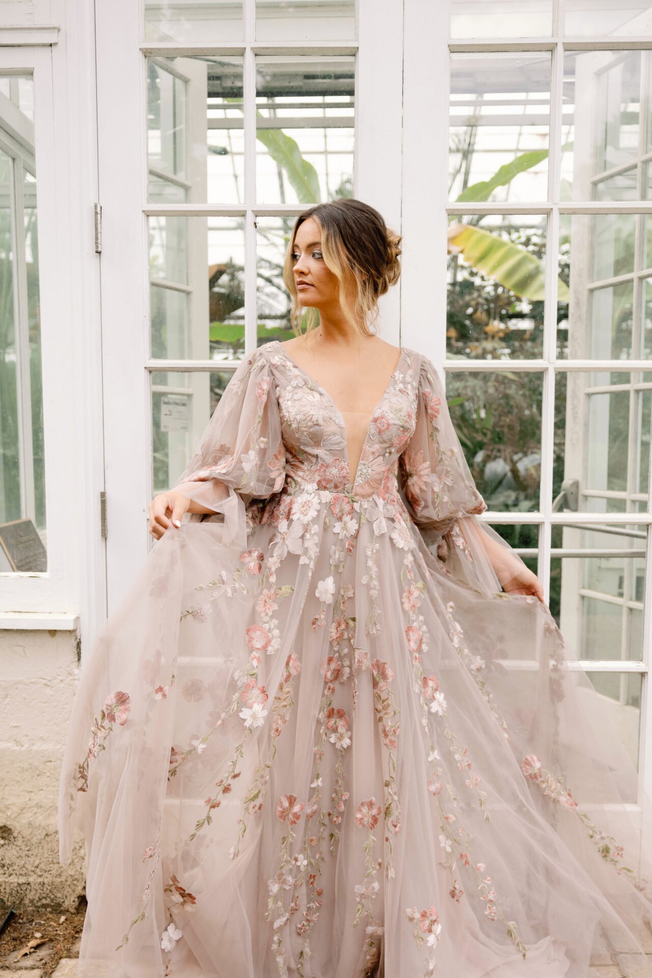 A woman in a long, embroidered, pastel-colored dress with floral details stands in front of a glass greenhouse door.