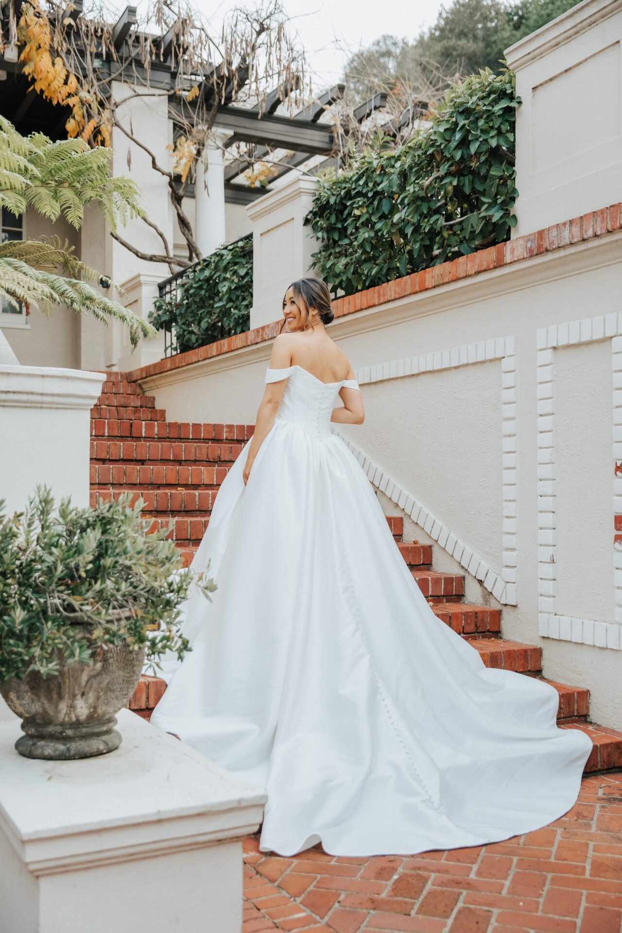 A woman in a white off-the-shoulder wedding gown smiling and turning over her shoulder on a brick staircase outside.