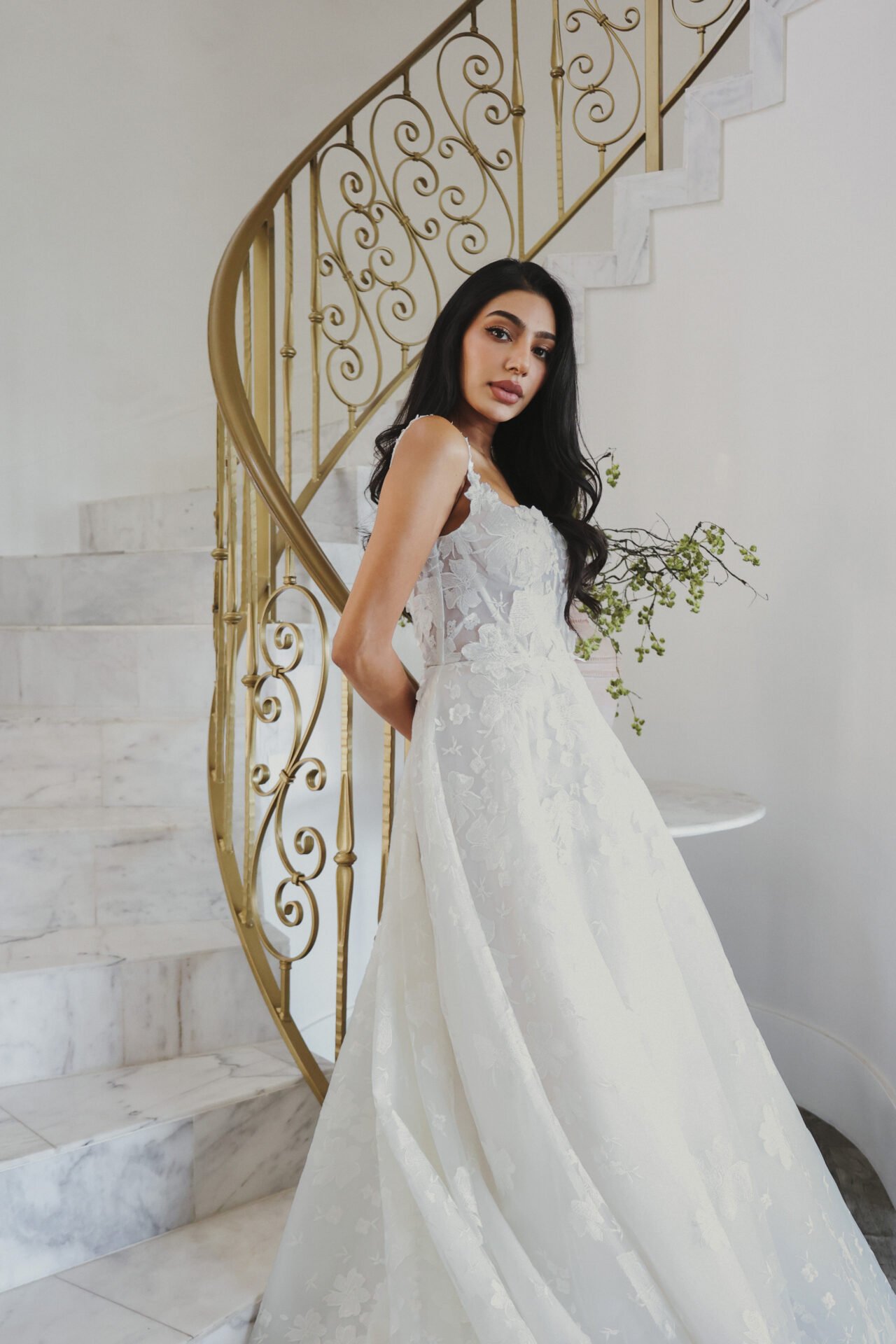 A woman in a white wedding dress with floral embroidery standing on a marble staircase with a gold railing.