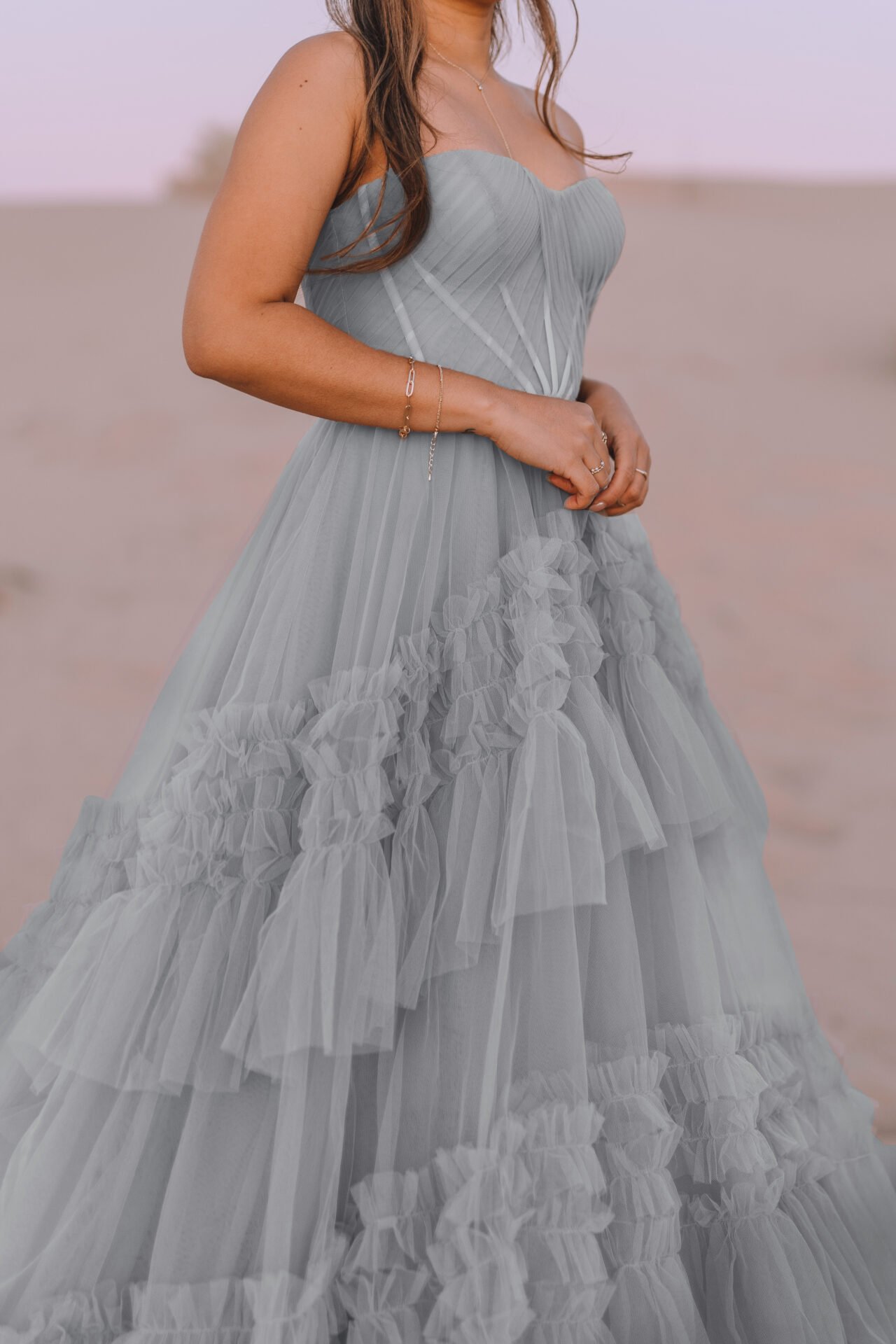 A woman wearing a strapless, light gray ruffled gown with pleated bodice standing outdoors in a desert landscape during sunset.