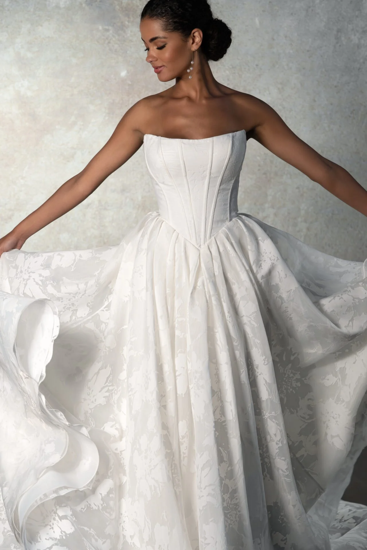 Woman in a strapless, white wedding gown with a full, textured skirt and corset-style bodice, holding out the fabric with her arms resting outward.