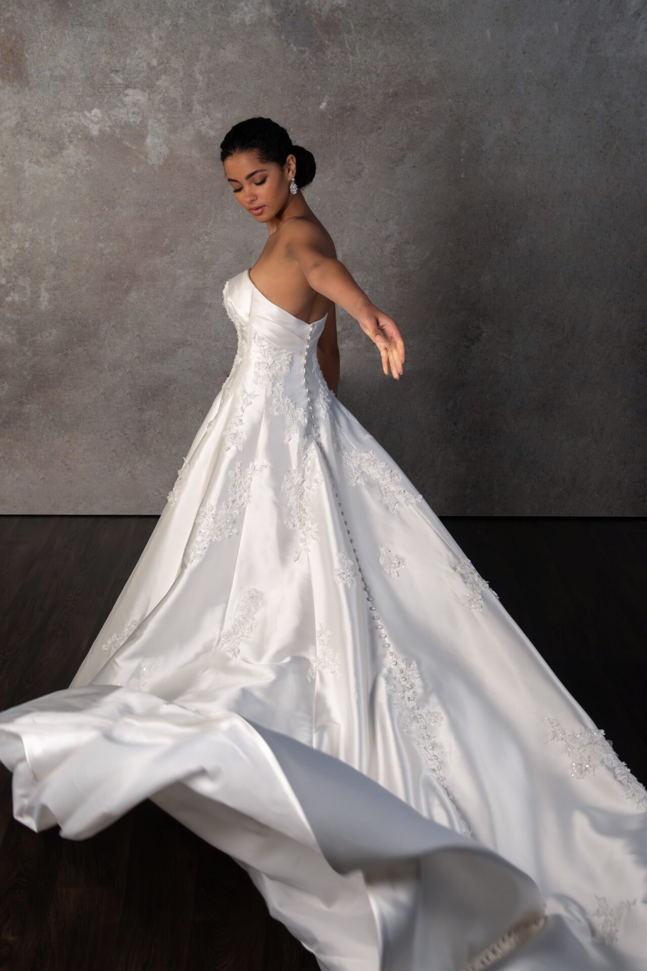 A woman in a white wedding gown with lace details dancing or posing in front of a textured gray wall.