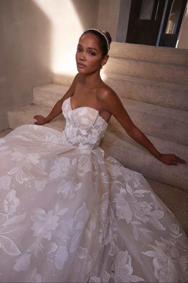 A woman wearing a strapless white gown with floral lace patterns, sitting on stairs with a neutral expression and natural makeup.