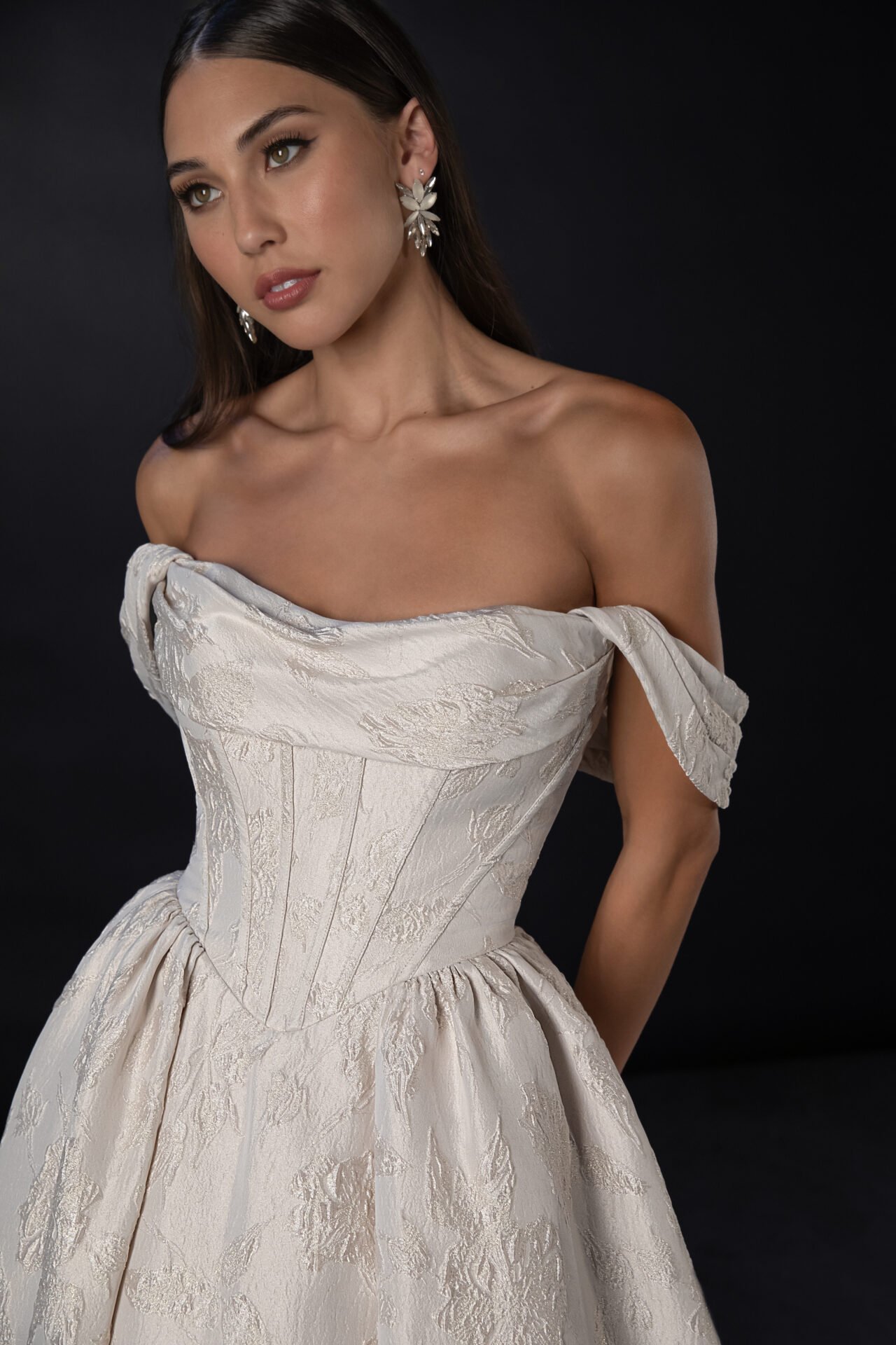 A woman in an off-shoulder, cream-colored, textured dress with flower-shaped jewelry against a dark background.