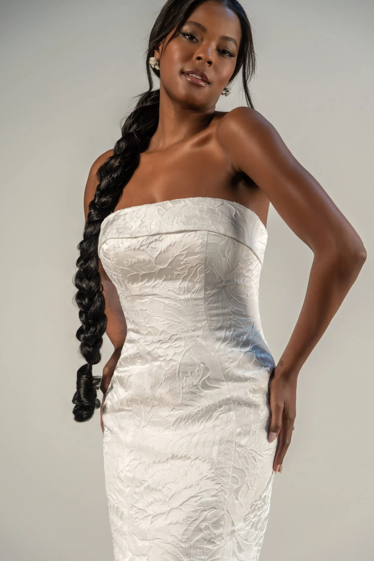 A woman with dark skin wearing a strapless white textured dress, posing with one hand on her hip, long black braided hair, and floral earrings.