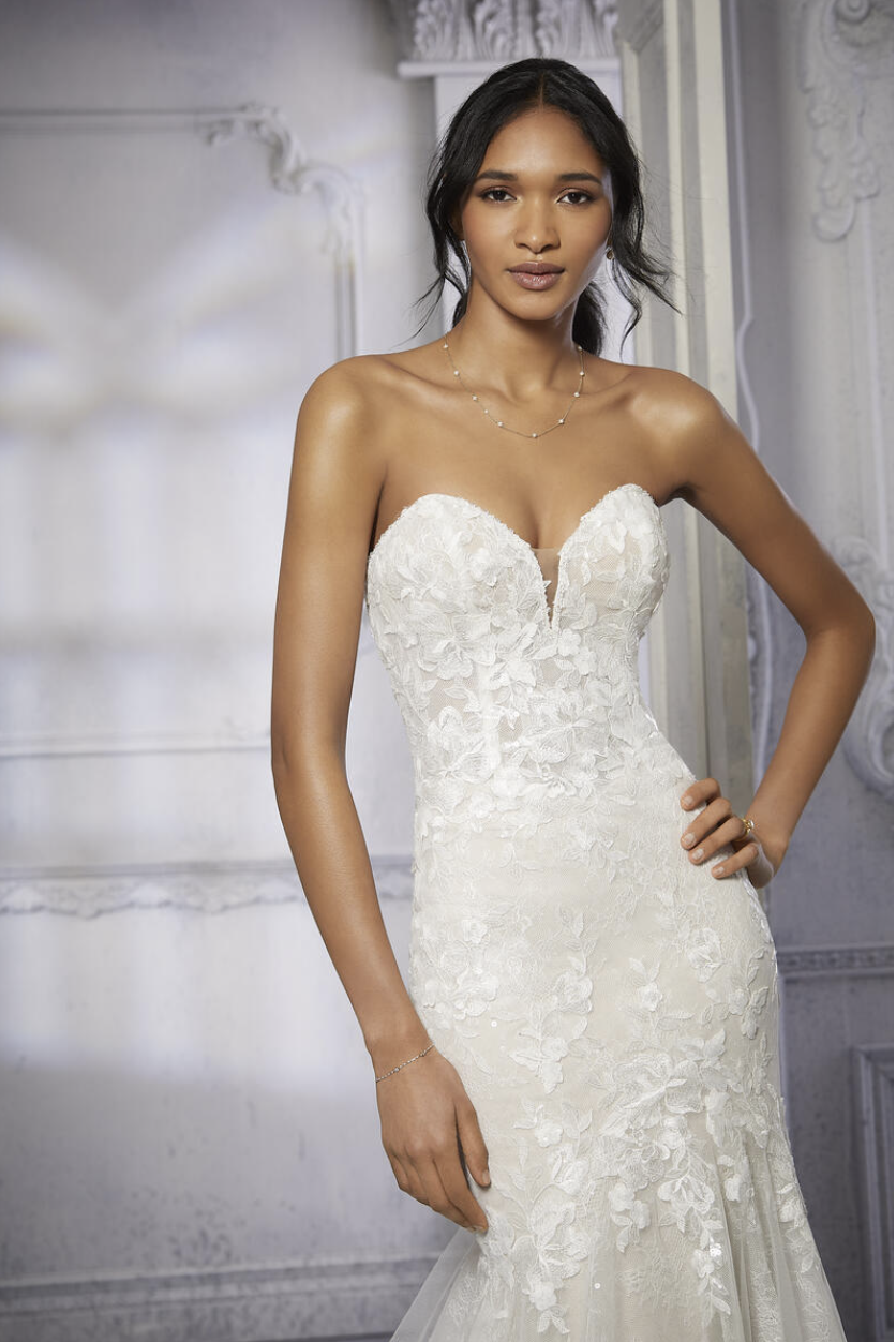 A woman in a strapless white wedding dress with floral lace details, standing indoors in front of a decorative wall.