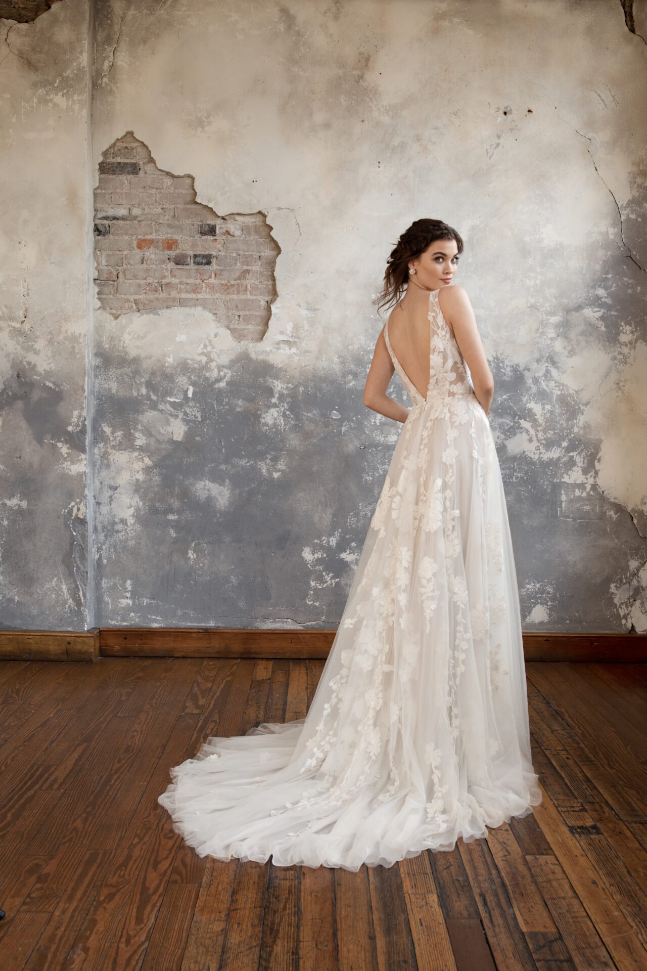 A woman in a white, floral-embroidered wedding dress with a deep V back, standing on a wooden floor against a weathered wall with exposed brick and peeling paint. She is looking over her shoulder.