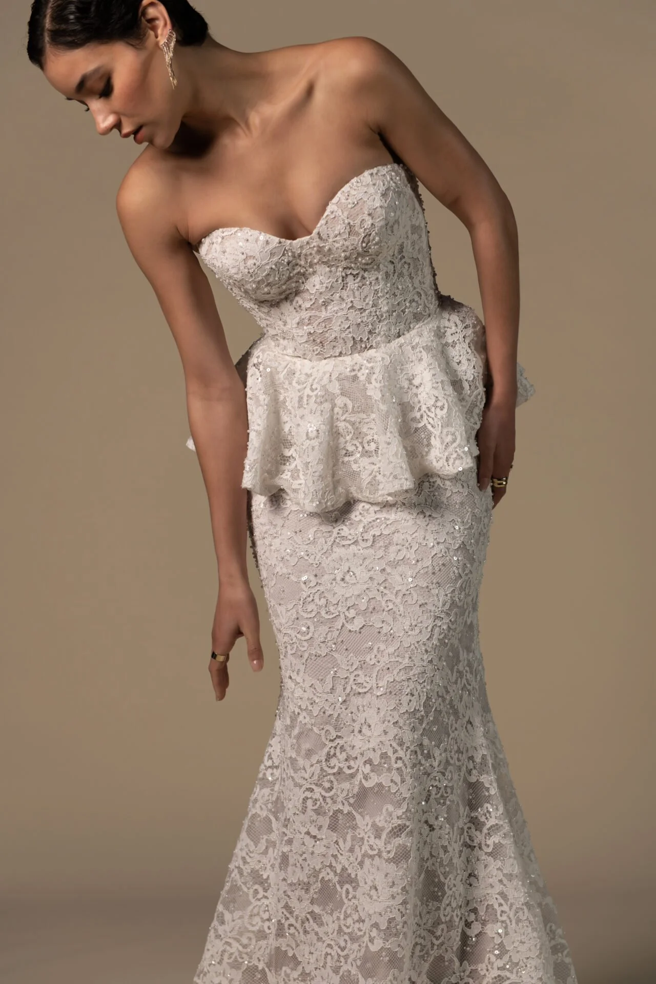 A woman wearing an elegant, strapless, white lace wedding dress with a peplum detail and a fitted, mermaid-style skirt. She has dark hair styled back and is wearing earrings.