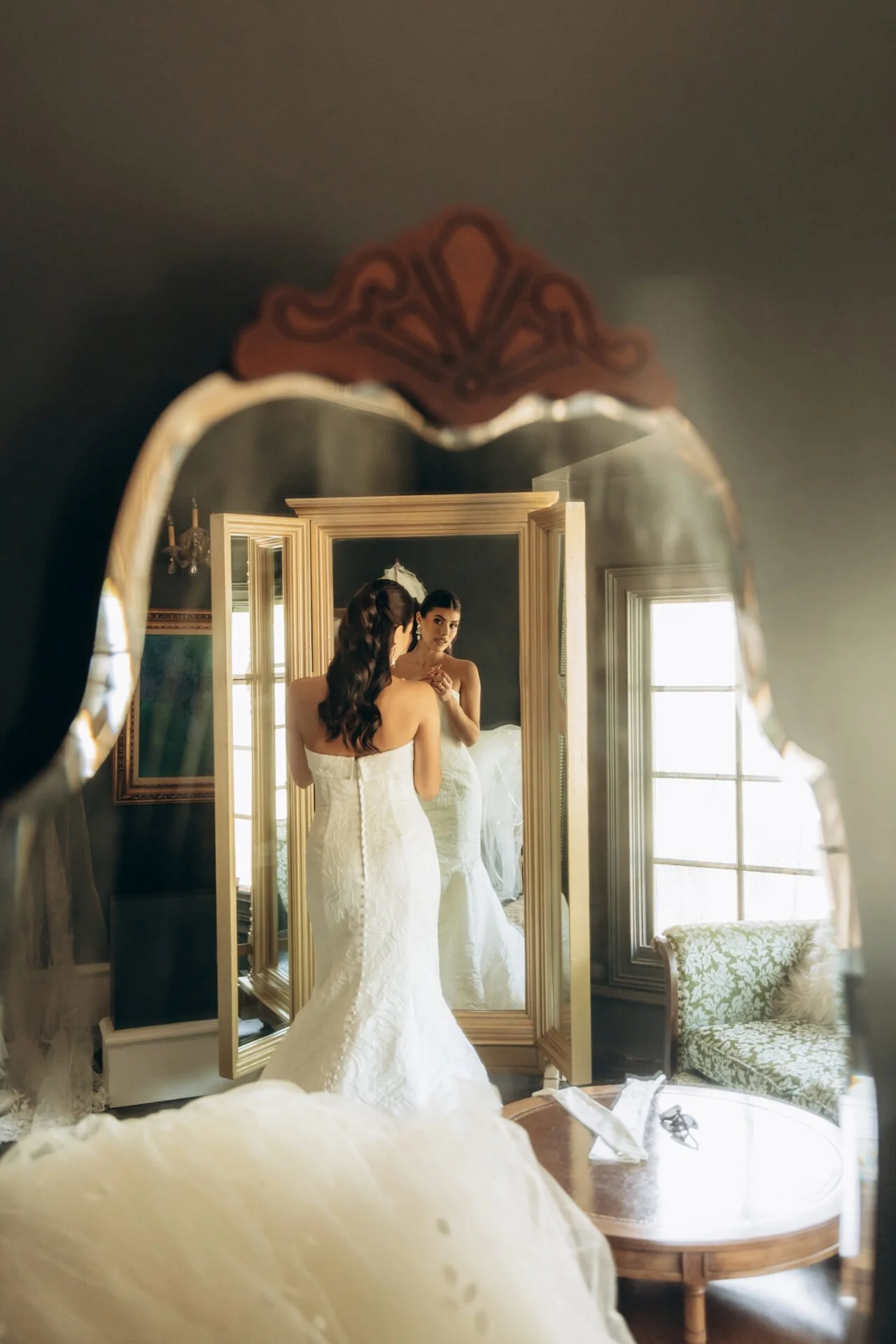 Bride in a white wedding dress looking at herself in a mirror in a vintage-style room with large windows.