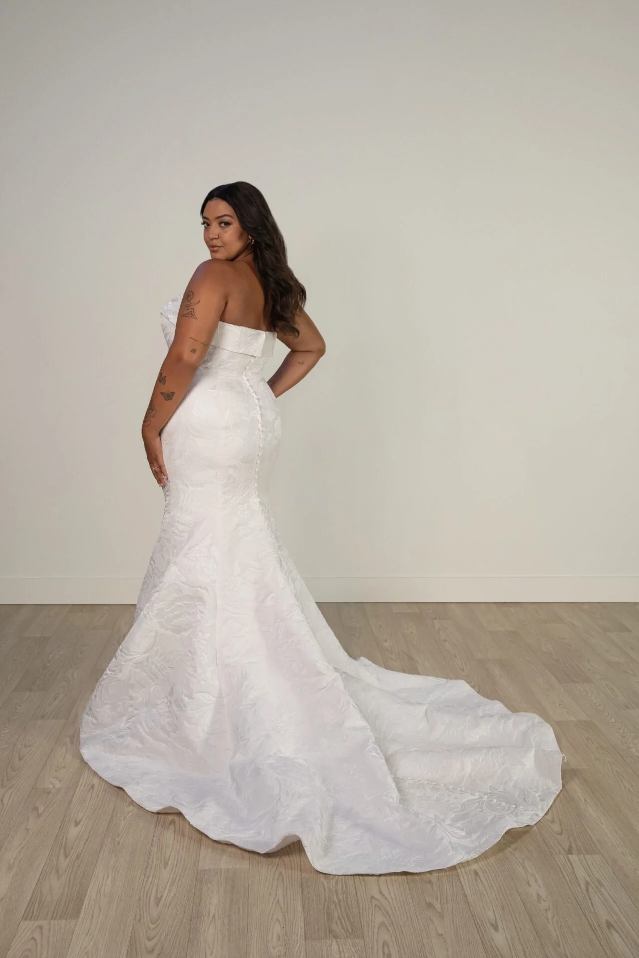 A woman in a strapless, white wedding gown with a train, standing on a wooden floor against a plain, light-colored wall