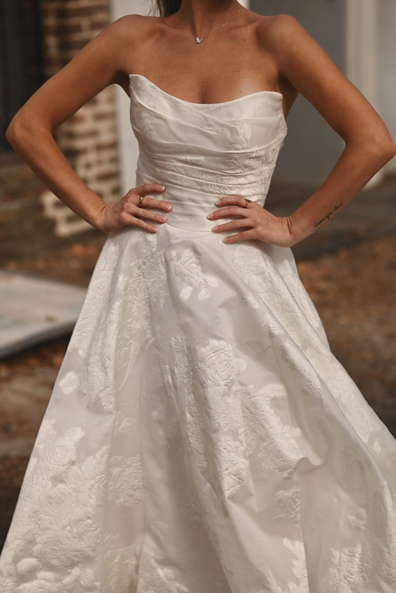 A woman wearing a white strapless wedding dress with floral embroidery standing outdoors with hands on hips.
