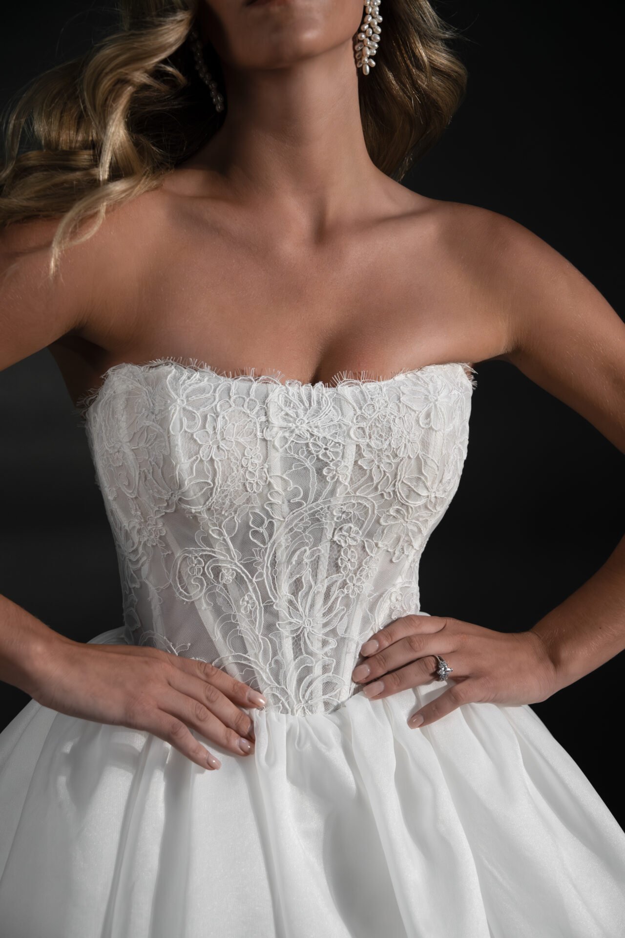 A woman wearing a strapless wedding dress with lace embroidery on the bodice, and large pearl earrings, with her hands on her hips, against a dark background.