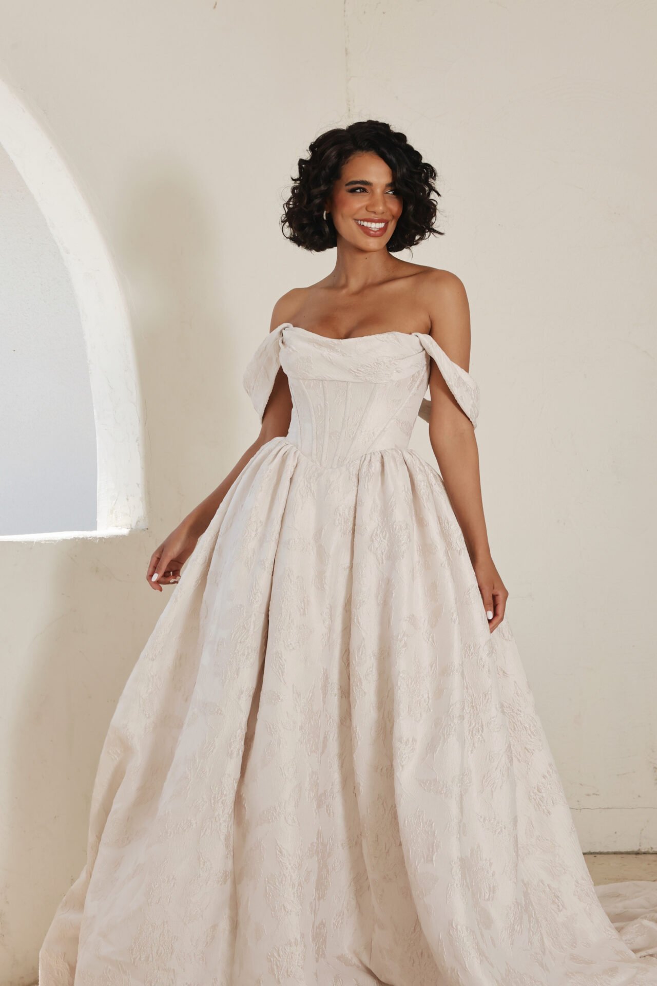 A woman in an off-shoulder, cream-colored formal gown with a textured pattern, standing in a minimally decorated room with an arched window.