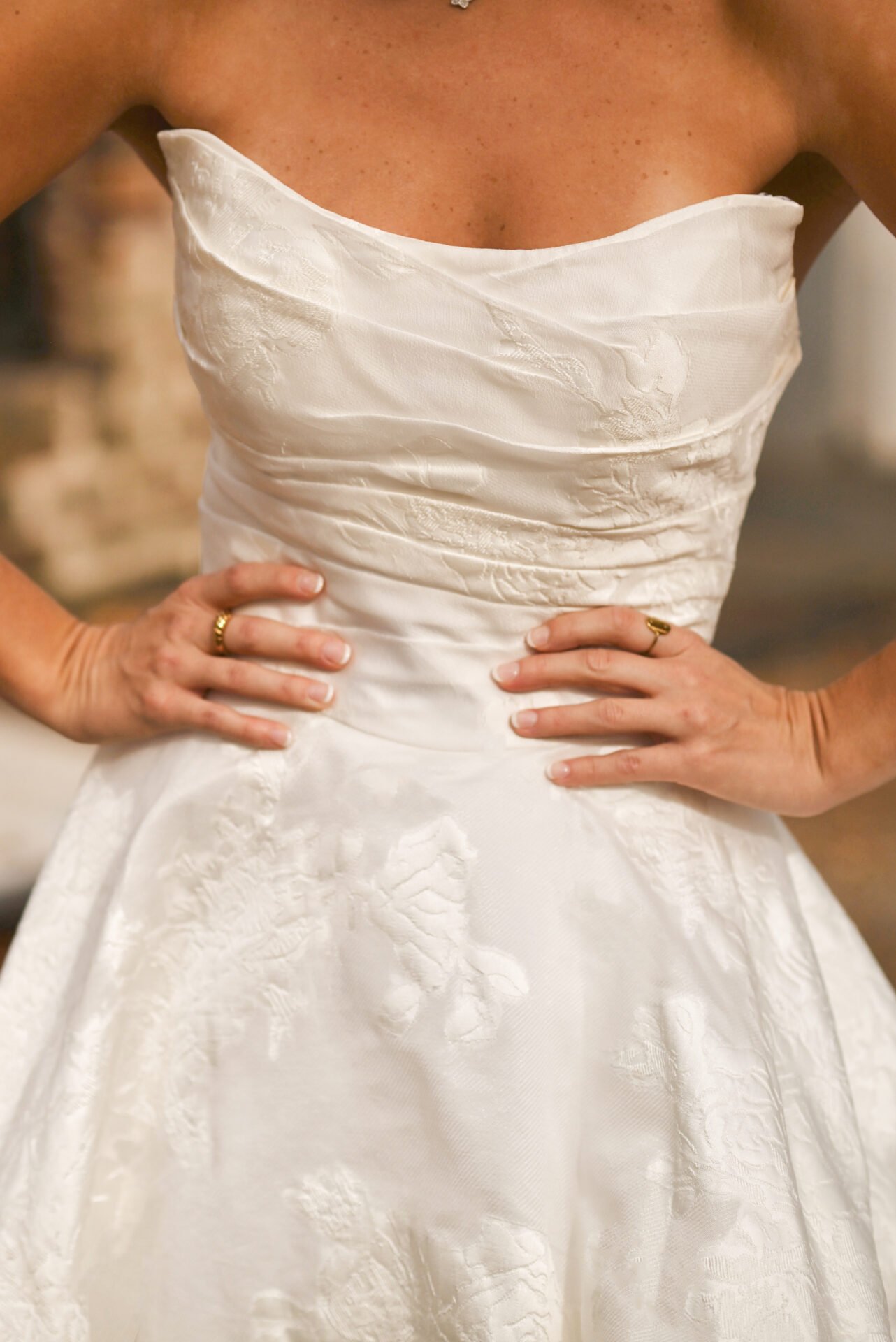 Close-up of a woman wearing a white strapless wedding dress with hands on hips, showing rings and textured floral fabric.
