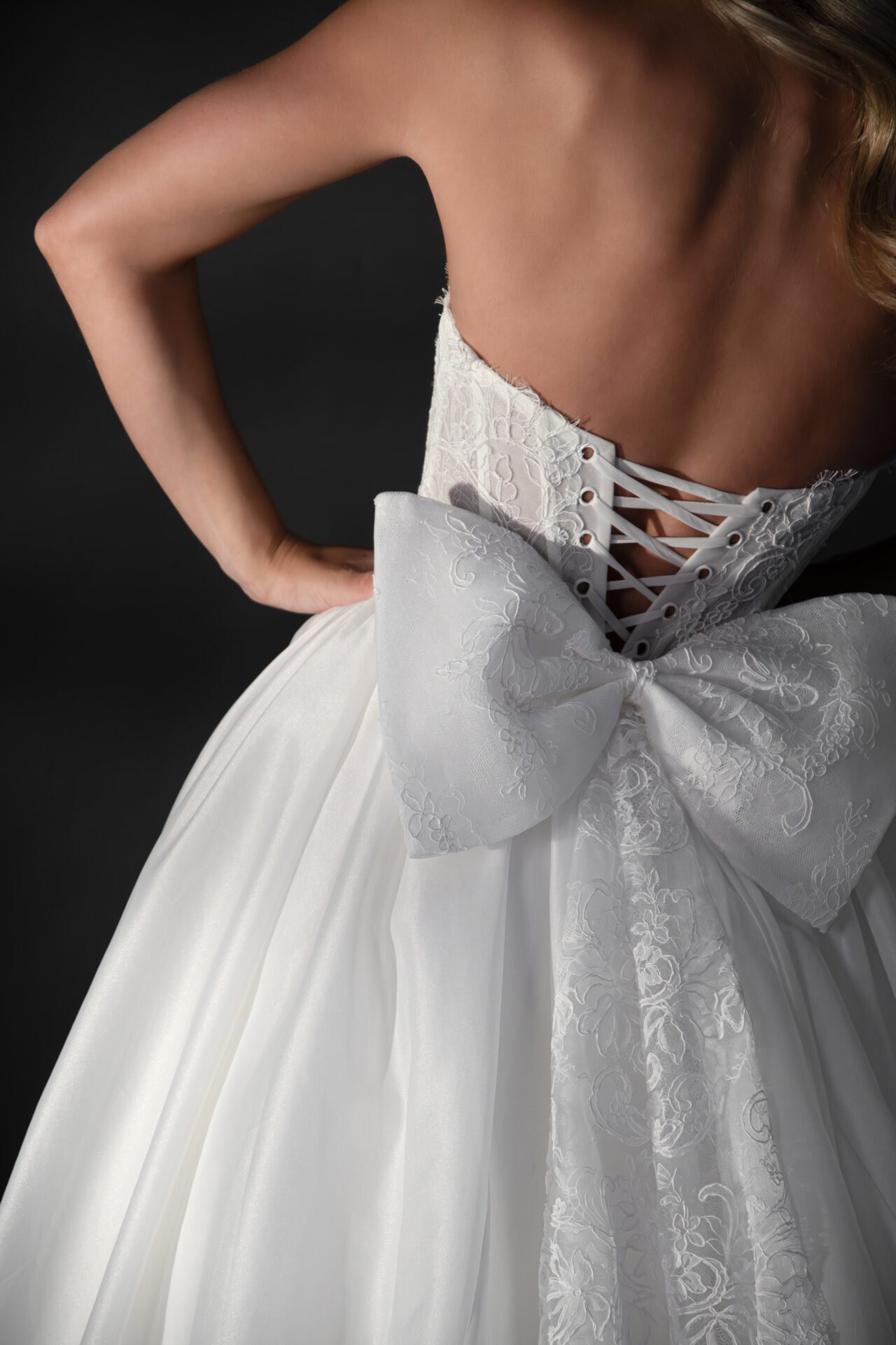 Close-up of a woman in a white wedding dress with a lace-up back and a large satin bow, standing against a black background.