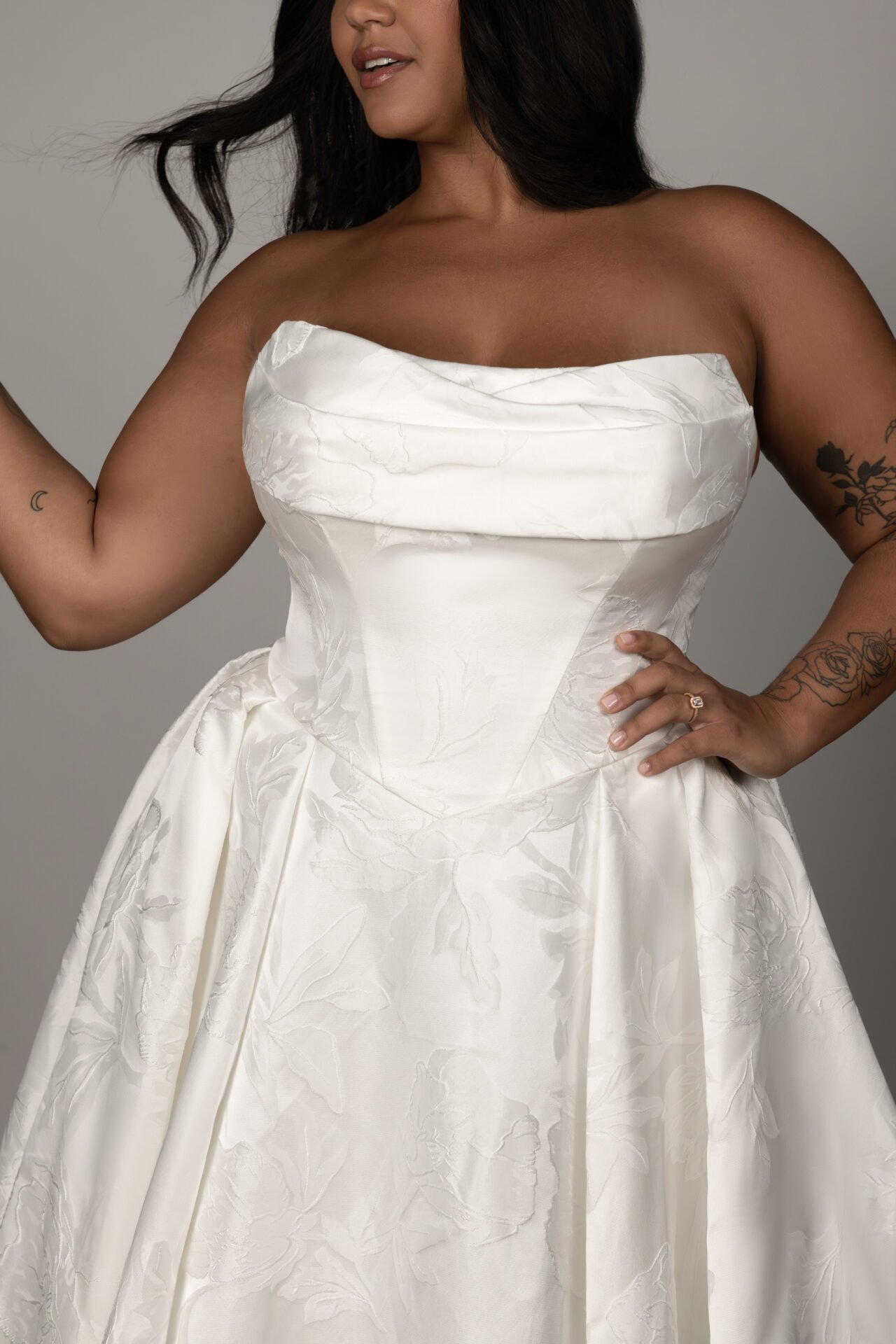 Woman wearing a strapless white wedding gown with floral embroidery, standing against a neutral background.