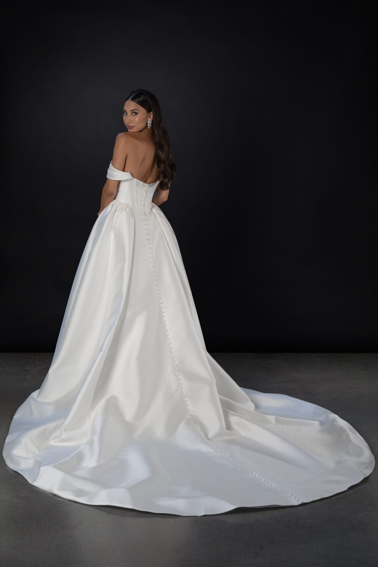 A woman in a strapless white wedding gown with a long train stands against a dark background, looking over her shoulder.