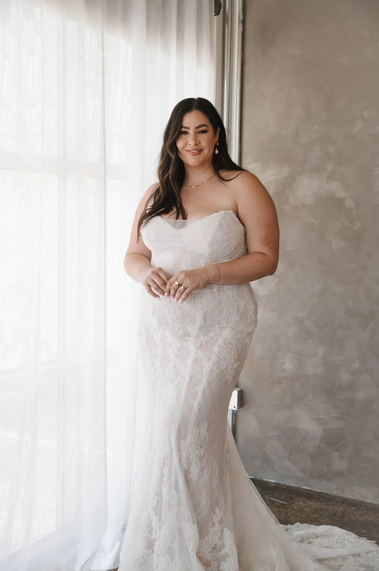 A woman in a white lace wedding dress standing indoors near a window with sheer curtains, smiling at the camera.