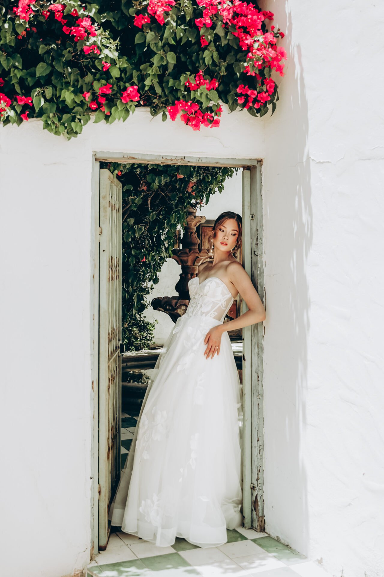 A woman in a white wedding dress standing in a doorway with greenery and pink flowers above, sunlight casting shadows on a white wall.