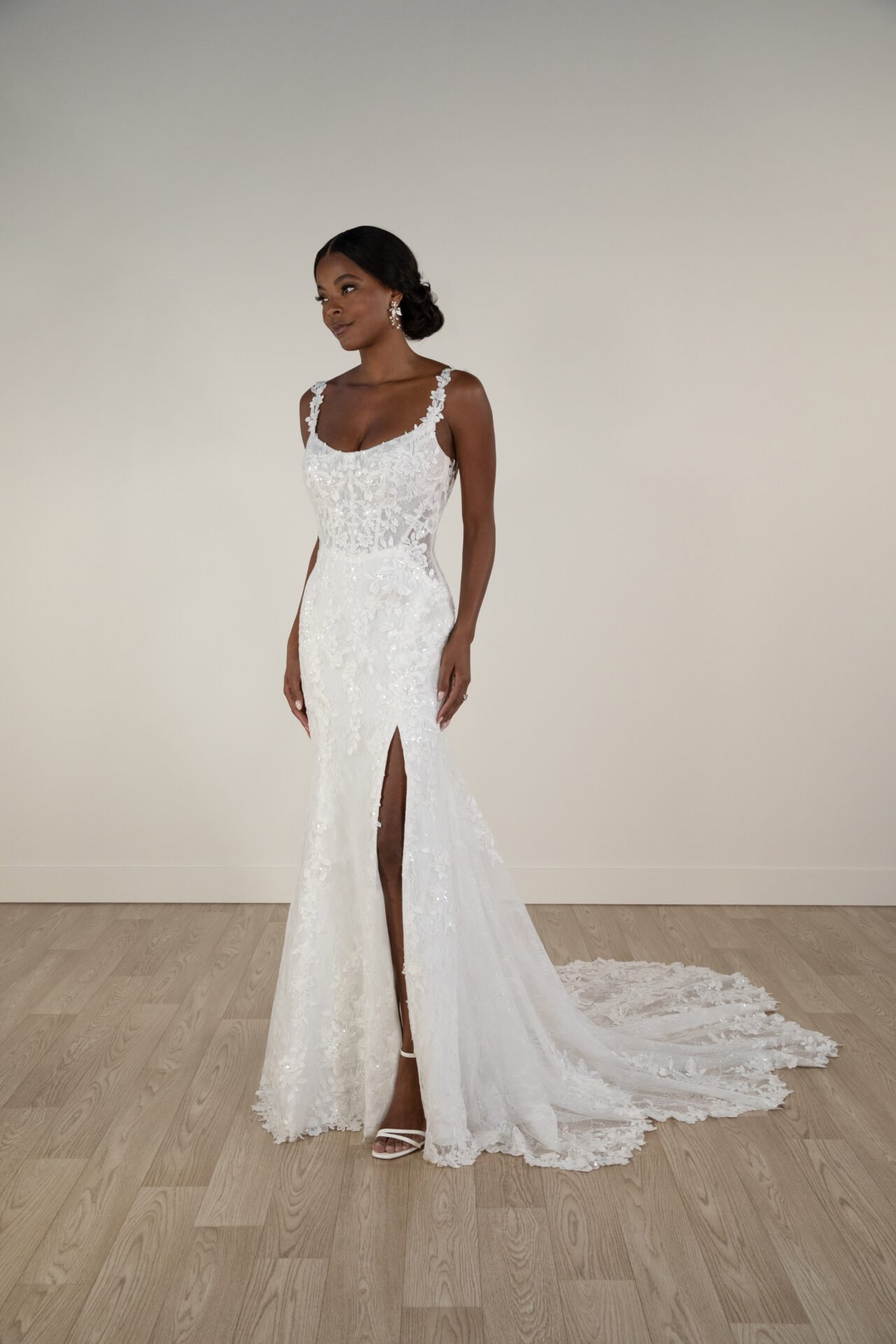 A woman in a white lace wedding dress with a high slit stands against a plain wall with wooden flooring.