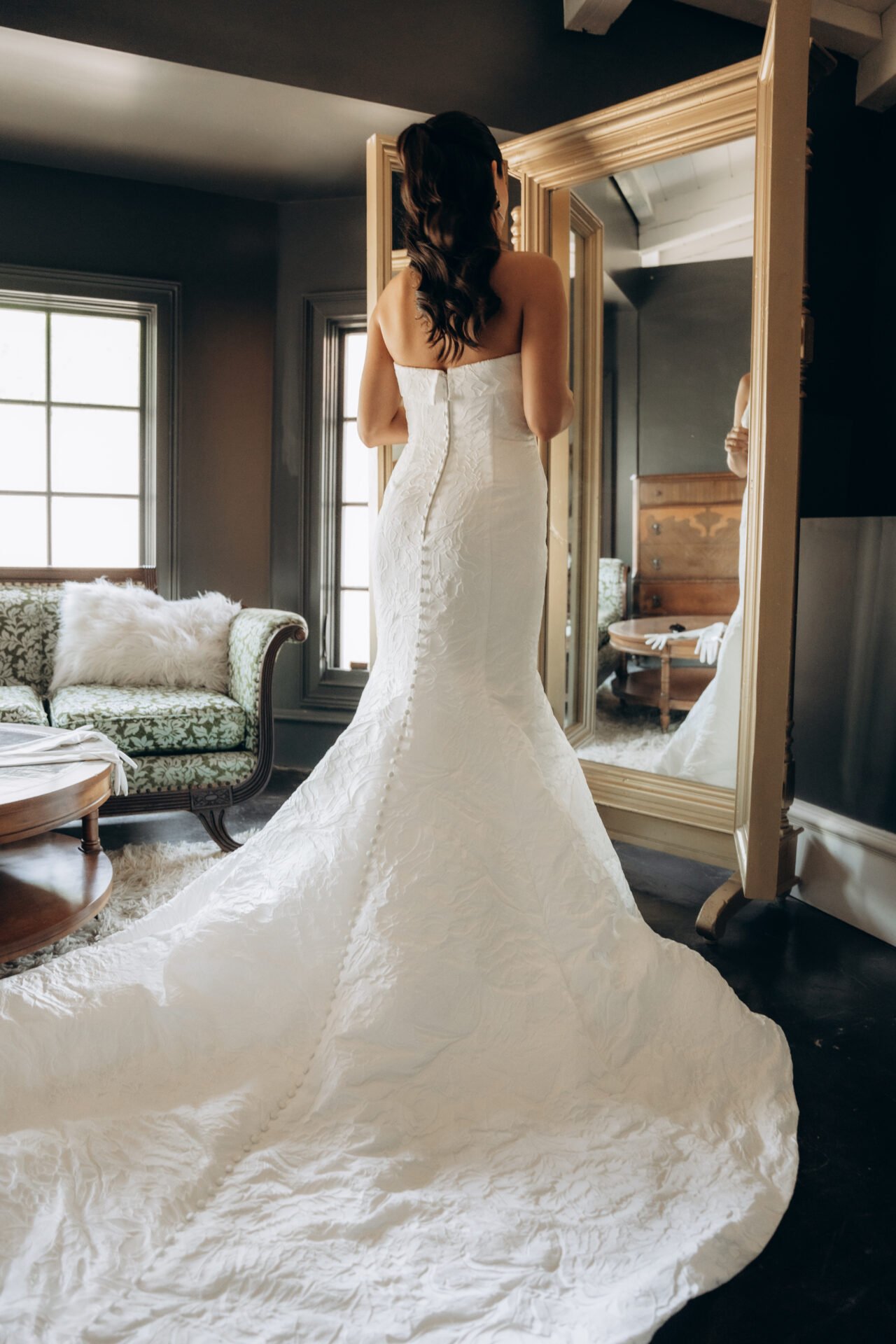 A woman in a strapless wedding dress looking at herself in a large mirror in a room with dark walls and two windows, with a vintage sofa and a wooden bench visible in the background.