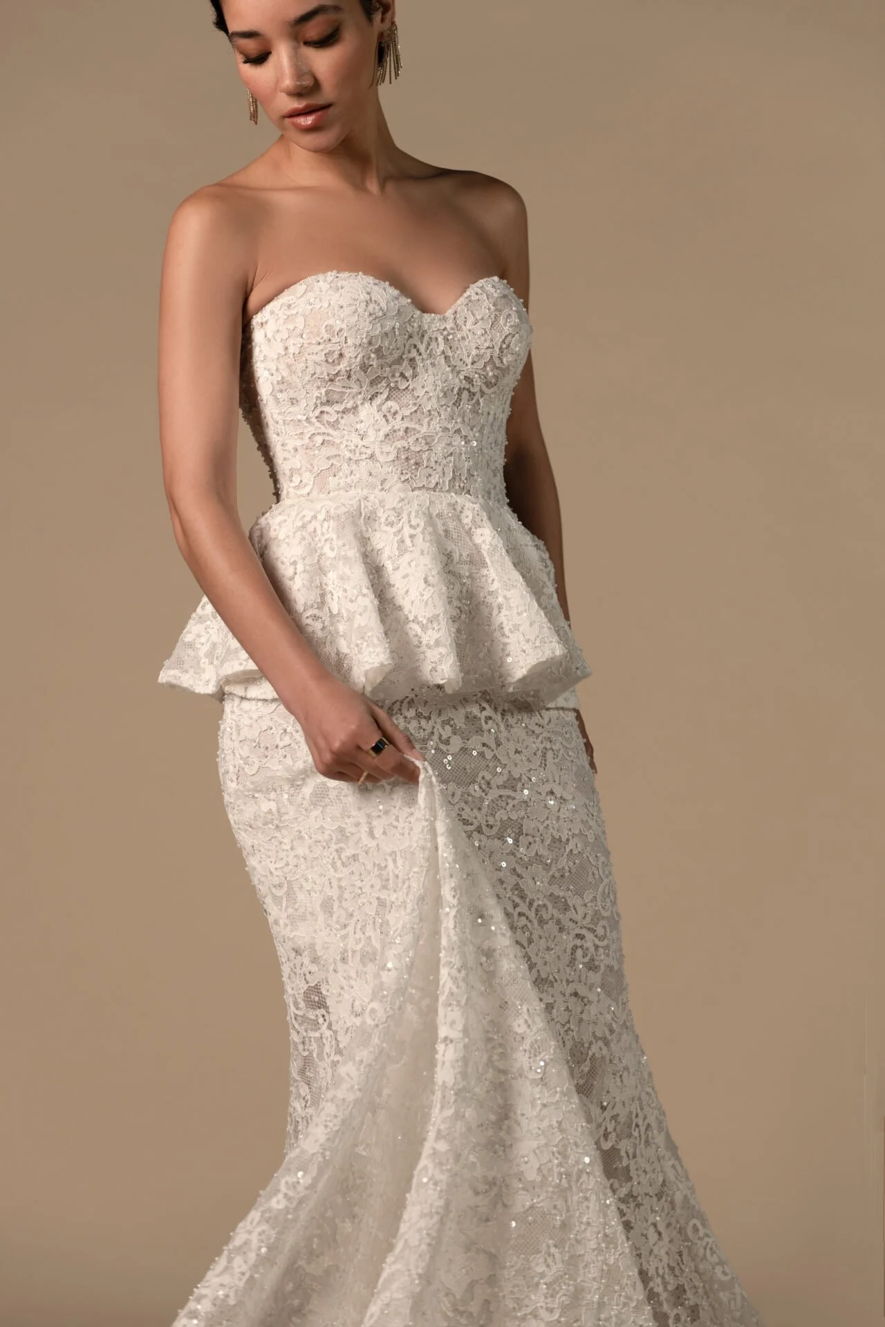 A woman wearing a strapless, lace wedding or evening dress with a ruffled waist and a long lace skirt, standing against a plain beige background.