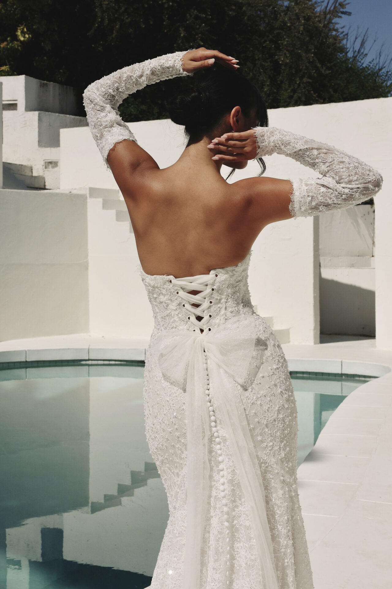 A woman in a white lace wedding dress with back lace-up and bow detail, wearing lace gloves, poses outdoors with arms raised and hands behind her head, near a pool with white Adirondack chairs and a white wall, under a clear blue sky.
