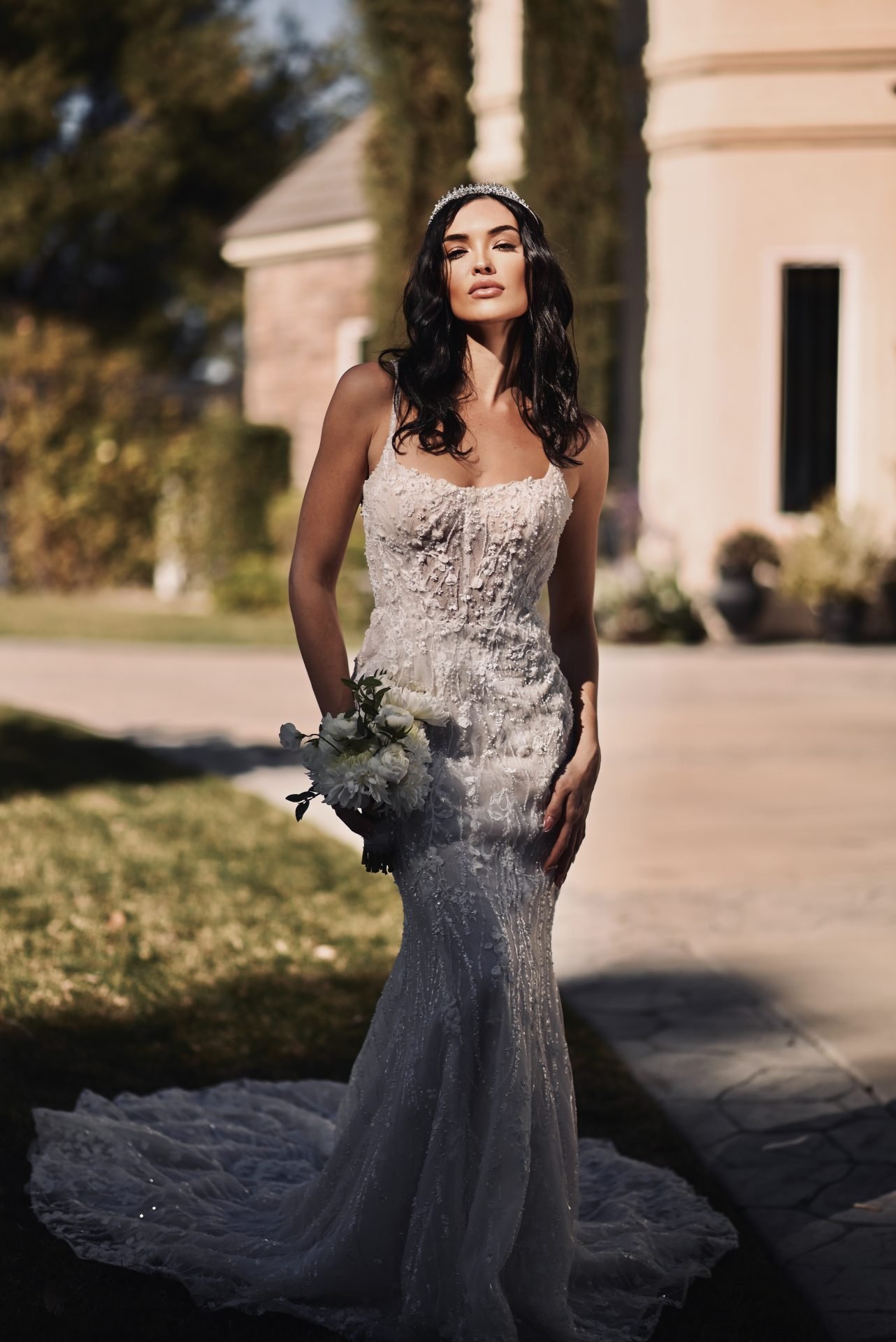 A woman in a white lace wedding dress holding a bouquet of white flowers outside on a sunny day.