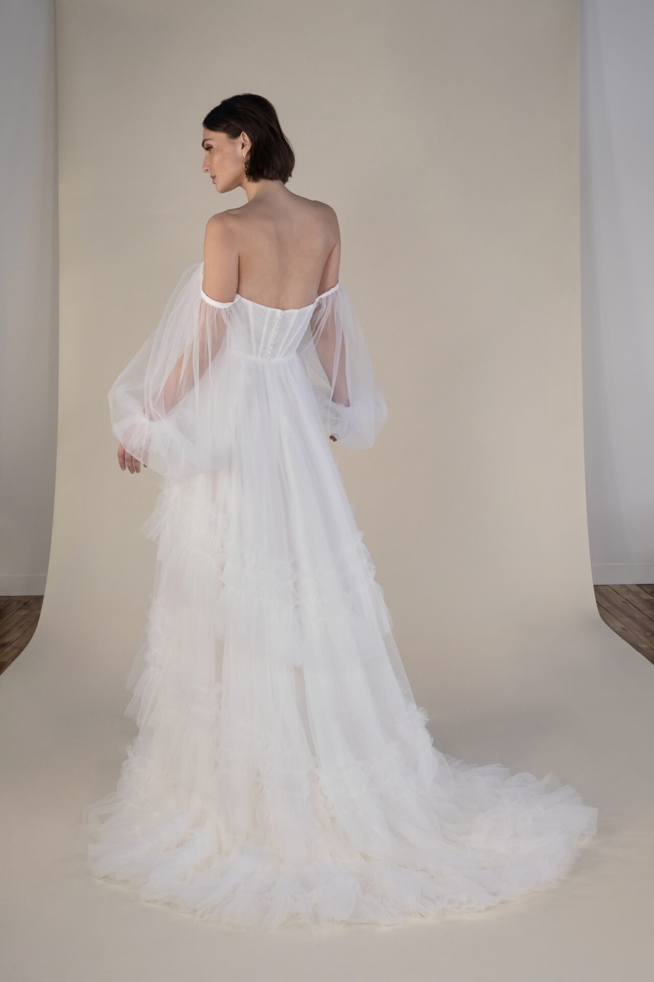 Woman in a white wedding dress with off-the-shoulder tulle sleeves, standing against a neutral background, shown from behind and in profile.