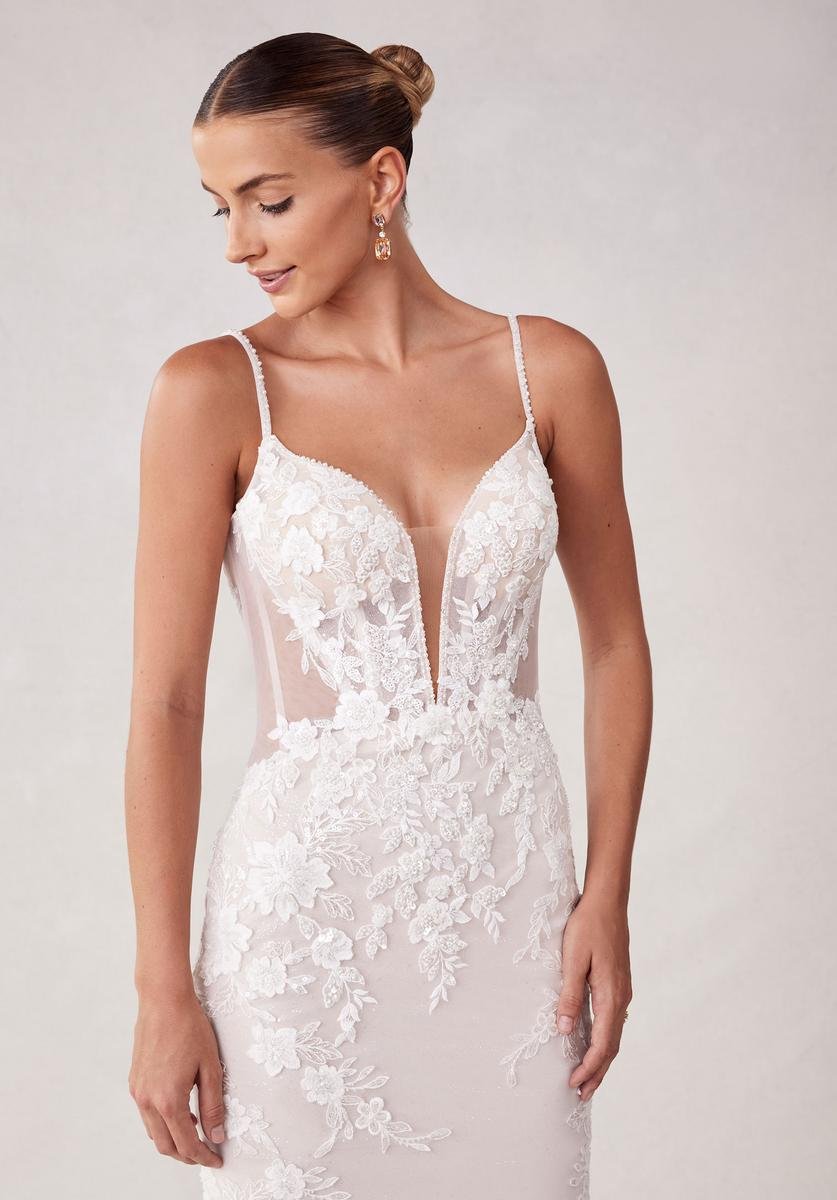 A woman wearing a white lace wedding dress with spaghetti straps and floral embroidery, looking down slightly, with her hair in a bun and wearing earrings.