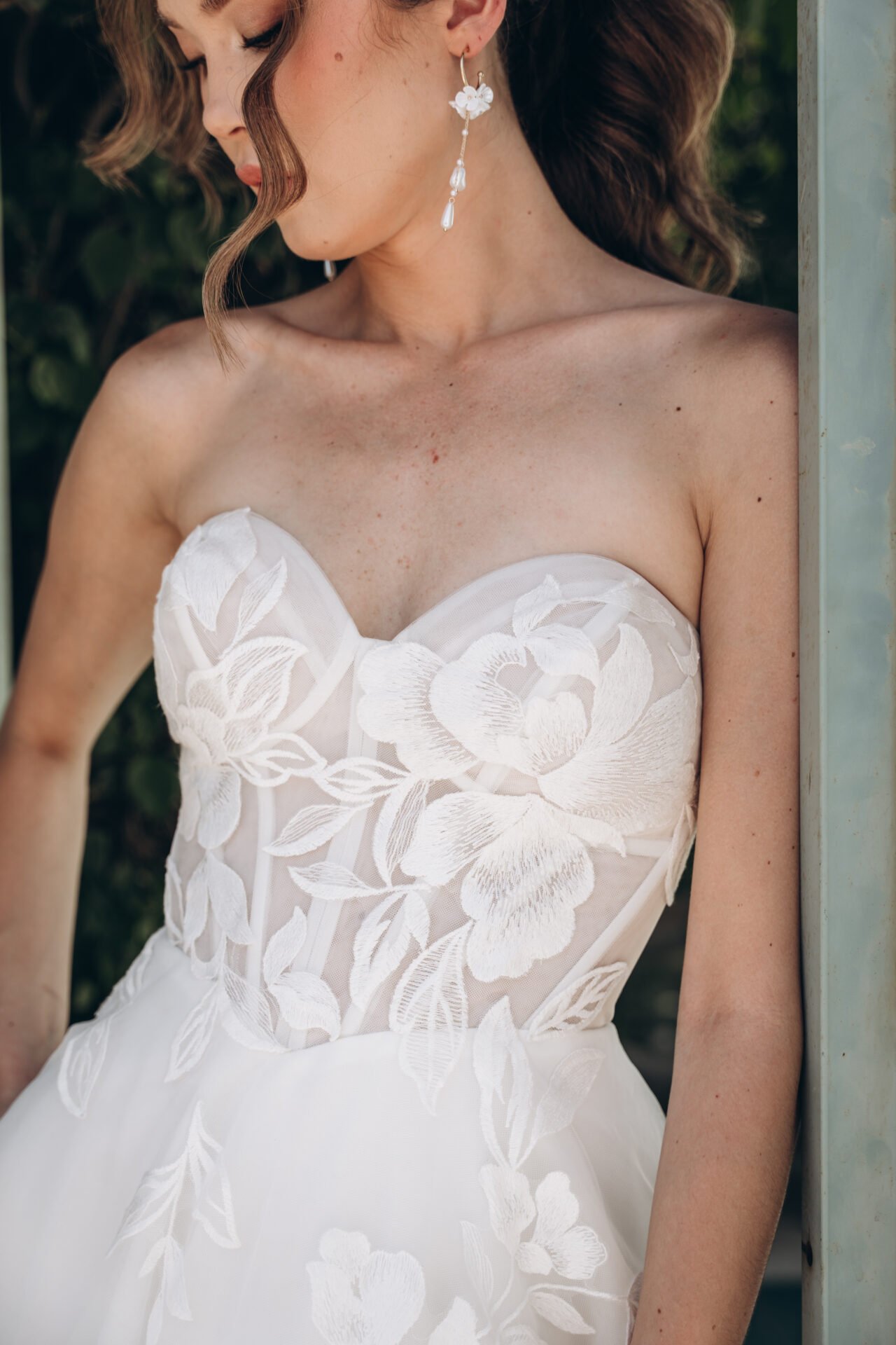 A woman in a strapless white wedding dress with floral embroidery, wearing floral drop earrings, standing near a metal post with greenery in the background.