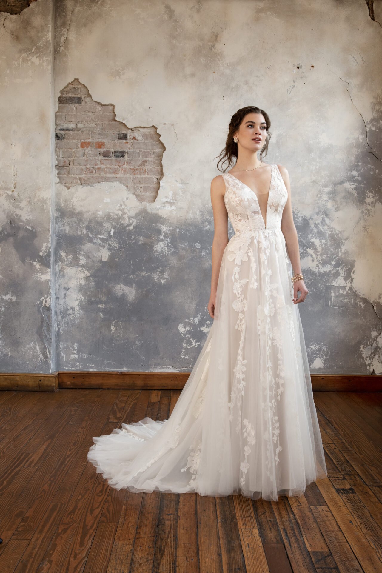 A bride in a cream-colored wedding dress with floral embroidery standing on a wooden floor in front of a distressed wall with peeling paint and exposed brick.
