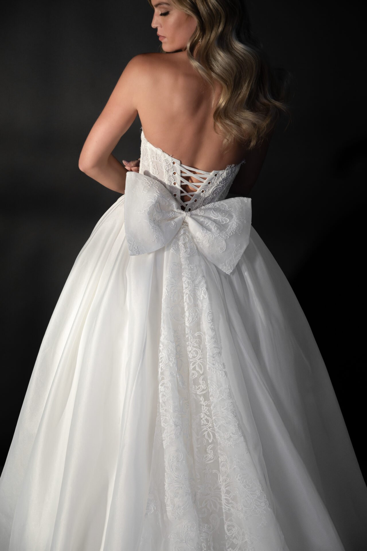A woman in a white wedding dress with a large bow at the back, lace-up corset detail, and flowing skirt, standing against a dark background.