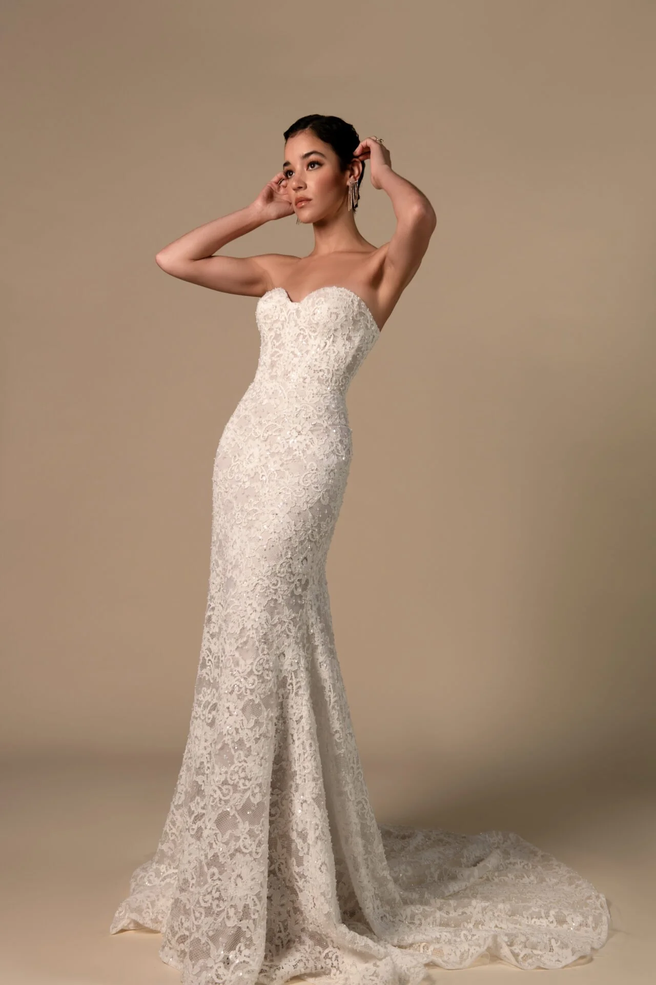 A woman wearing a strapless, fitted, lace wedding dress with a slight train, standing against a beige background with her hands touching her hair.