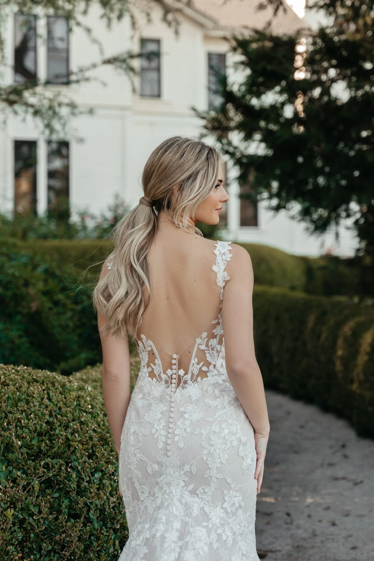 Woman in a white lace wedding dress with a low back and floral embroidery, standing outdoors near a hedge, with a white house and green trees in the background.