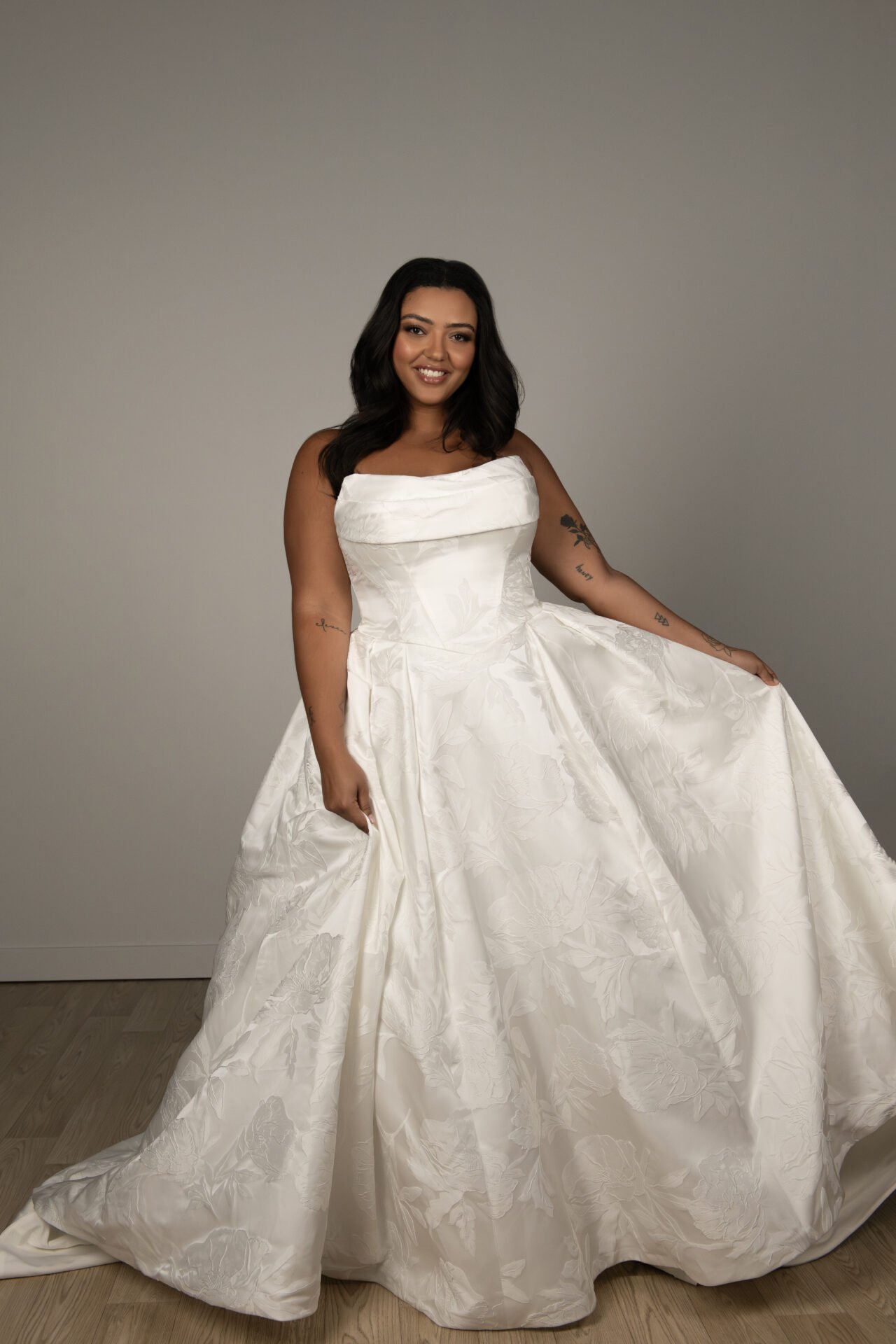A woman in a strapless white wedding gown with floral pattern, standing on a wooden floor against a plain wall, smiling and holding up part of her dress.