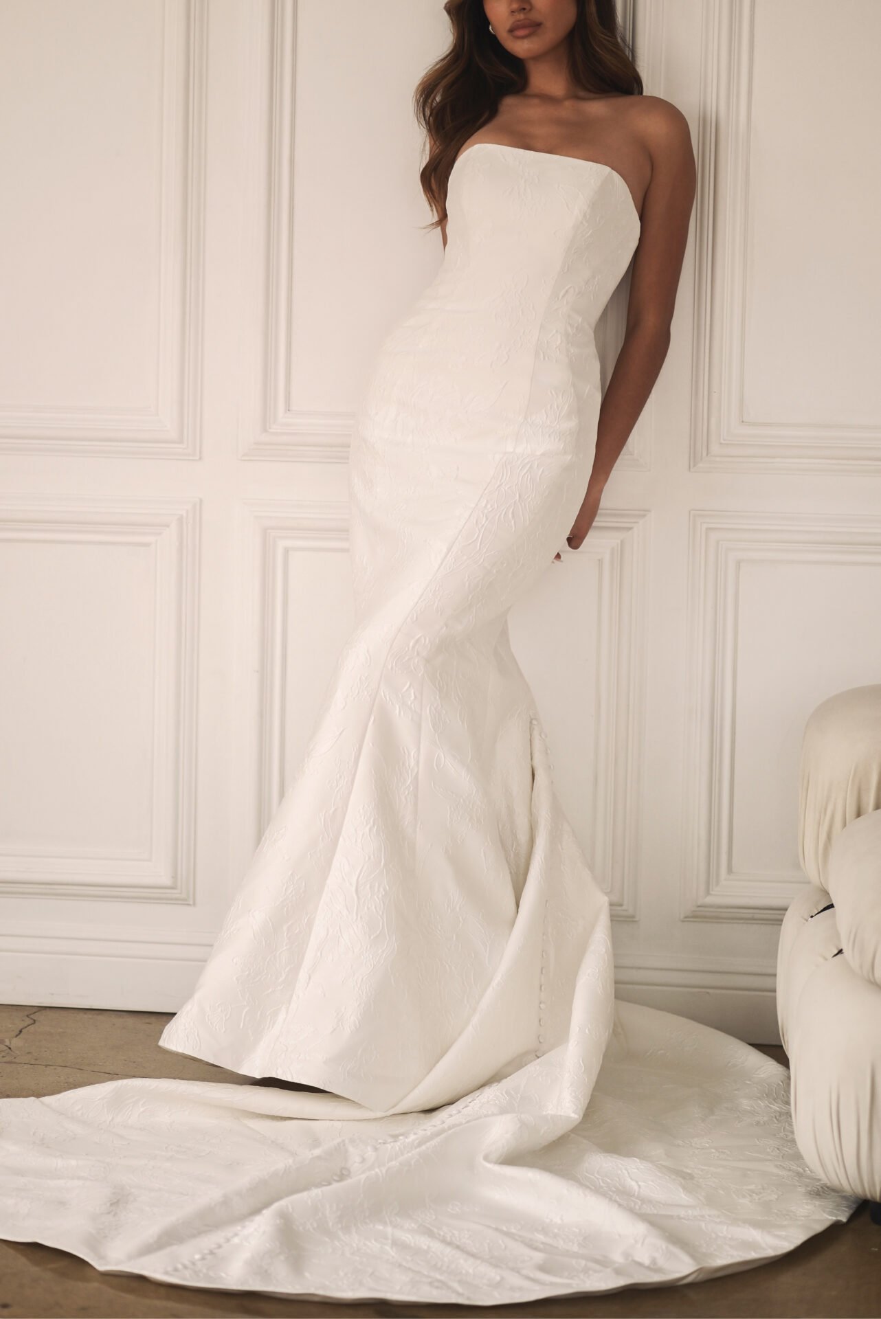 A woman is wearing a strapless, white wedding dress with a fitted bodice and a long train, standing in front of elegant white paneled walls.