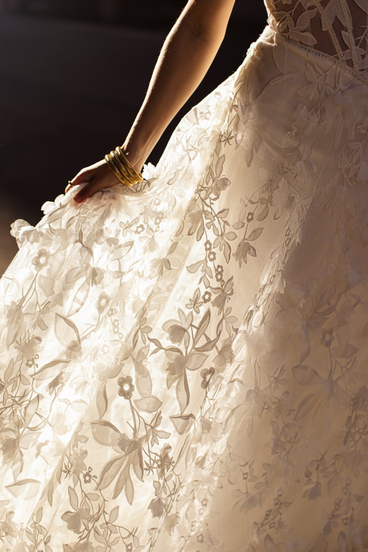 Close-up of a woman in a wedding dress with floral embroidery, holding the skirt and wearing multiple gold bangles on her wrist.
