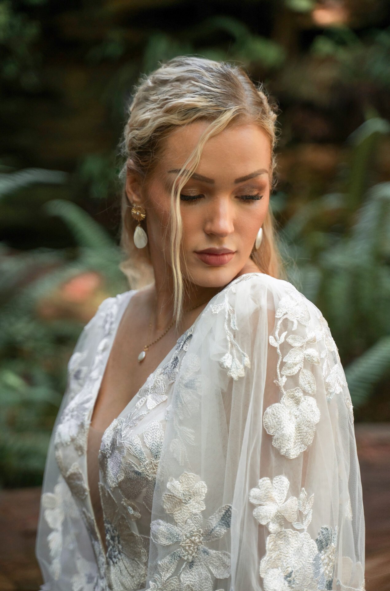 A woman with blonde hair, wearing a white embroidered dress and pearl jewelry, posed outdoor among green foliage.