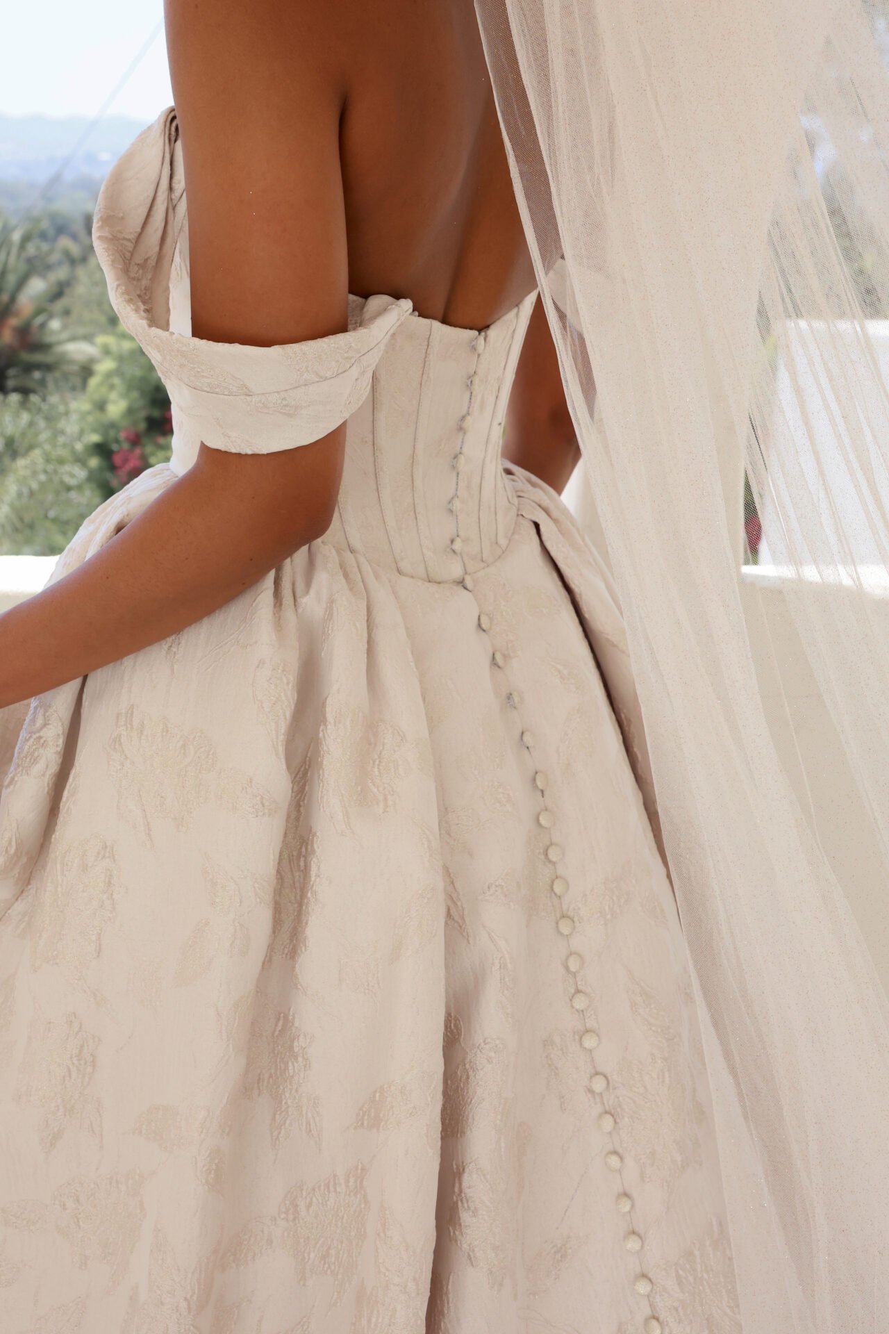 Close-up of a woman wearing a vintage cream-colored dress with a fitted bodice and button details, with a sheer curtain and outdoor greenery in the background.