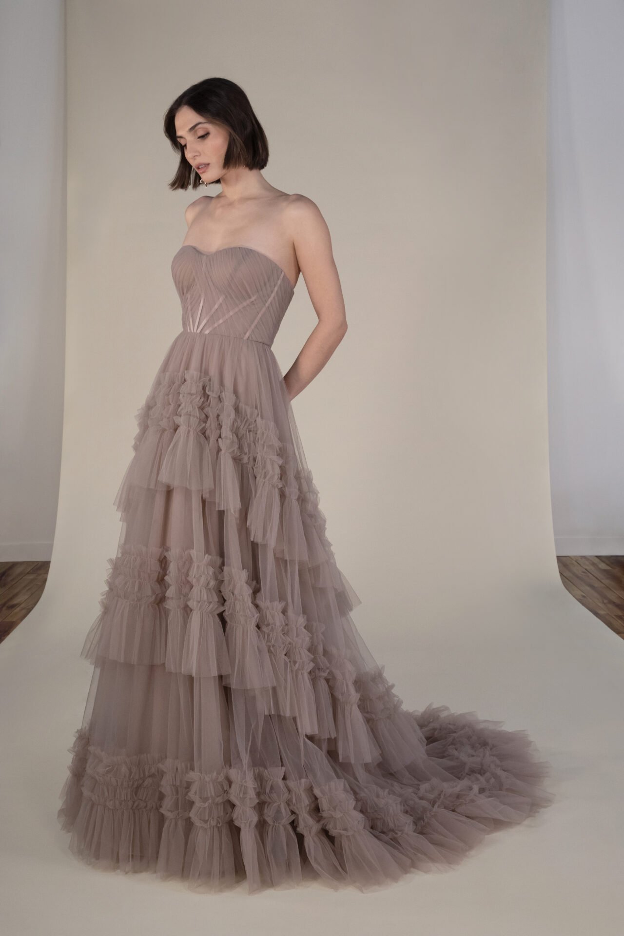 A woman in a strapless, long, ruffled, beige tulle dress stands on a white backdrop with her hands behind her back, looking down.