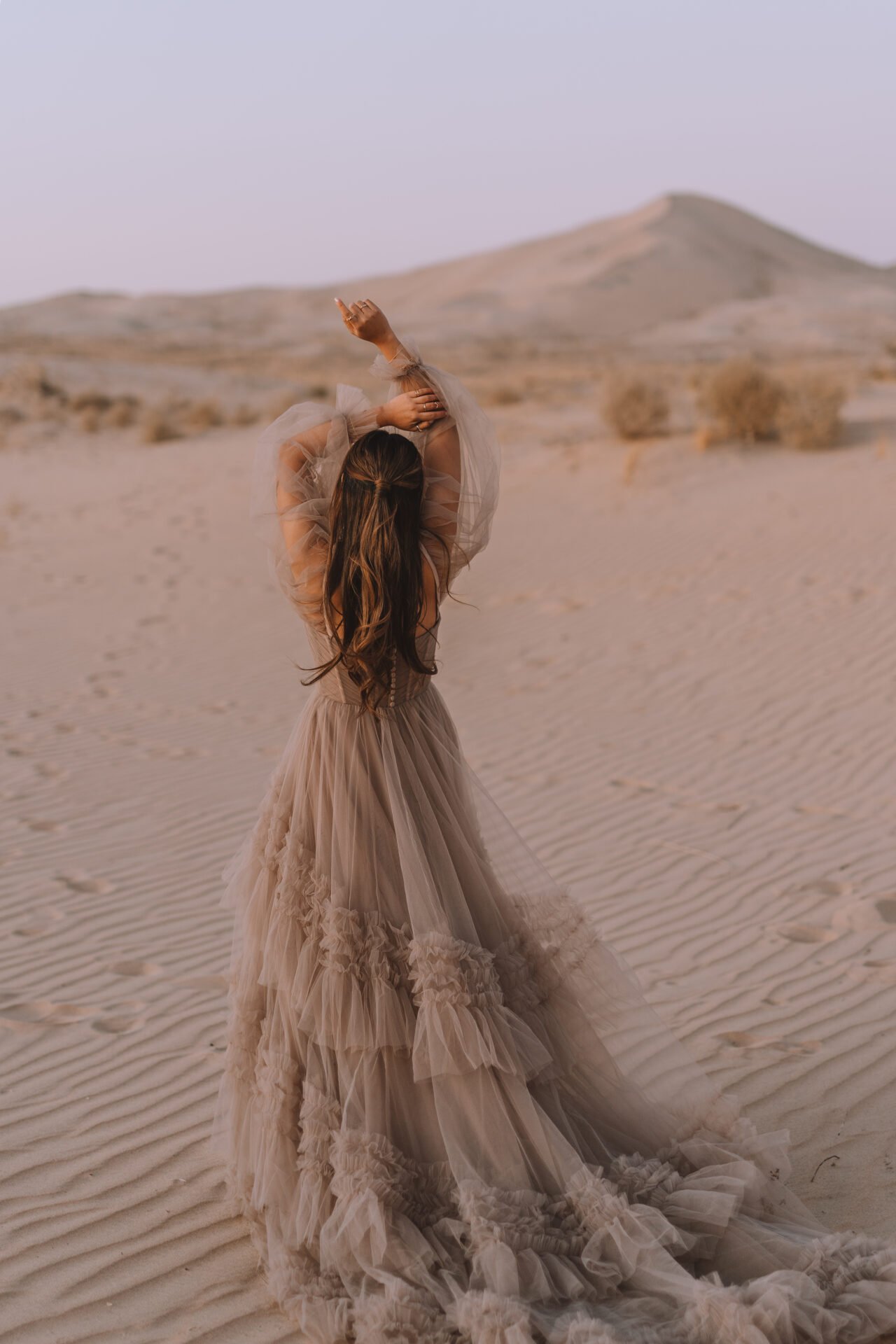 A woman in a flowing, beige tulle gown stands alone in a desert landscape with sand dunes and sparse vegetation, raising her arms and arched back under a pale sky.