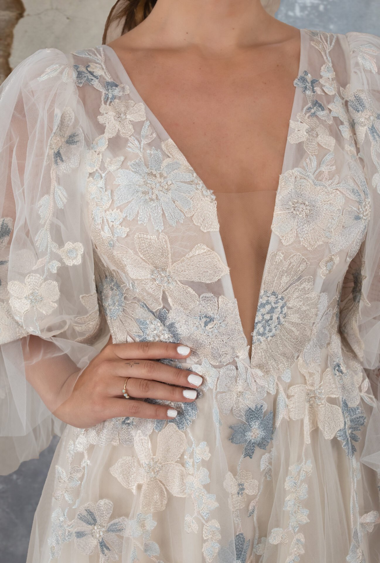 Close-up of a woman wearing an elegant, sheer, cream-colored dress with light blue and cream floral embroidery. The dress has a deep V neckline, and the woman's hand with white-painted nails and a delicate ring is resting on her hip.