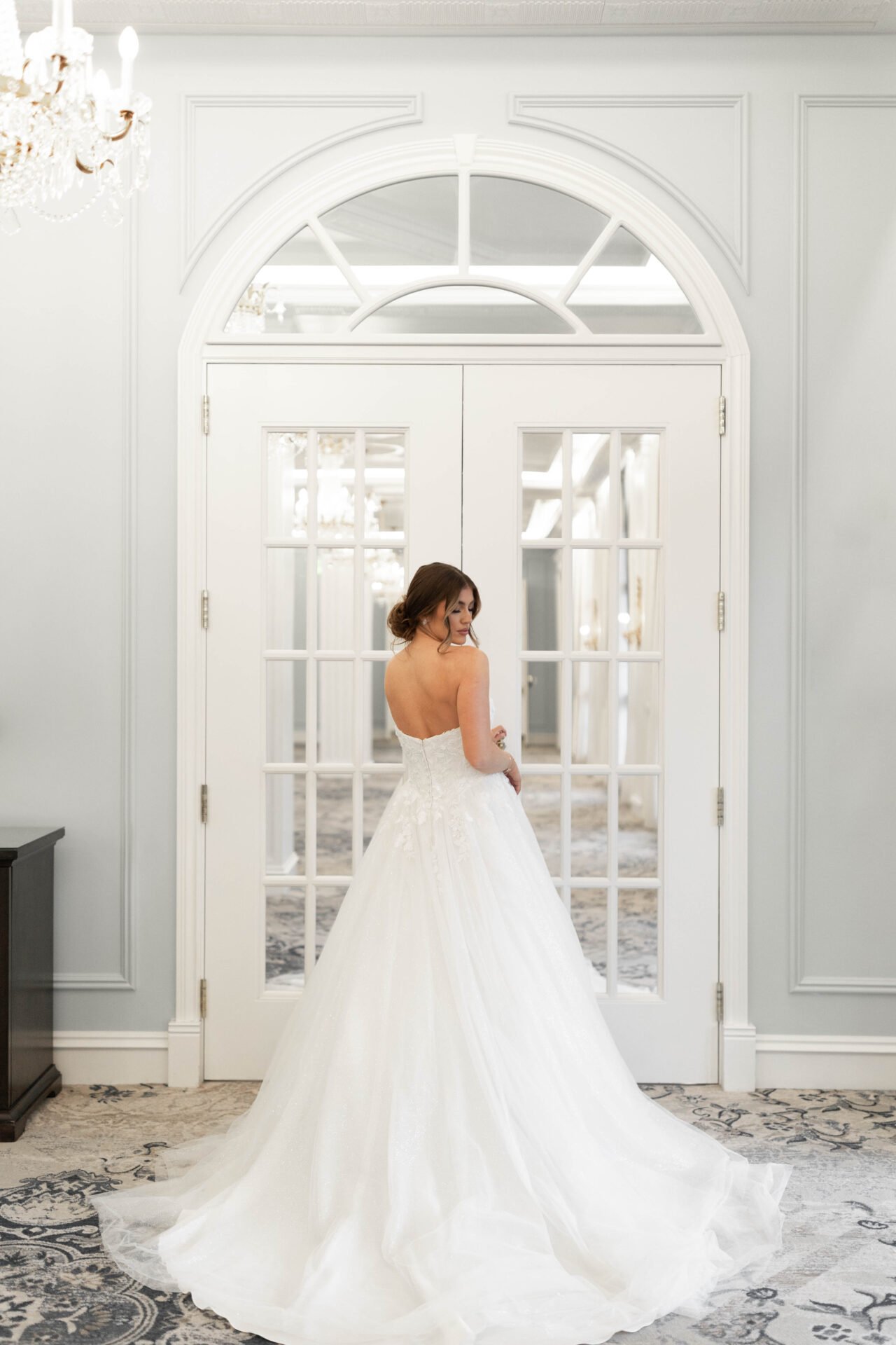 Brunette woman in a white strapless wedding gown stands in front of glass double doors in an elegant, bright room with chandeliers.