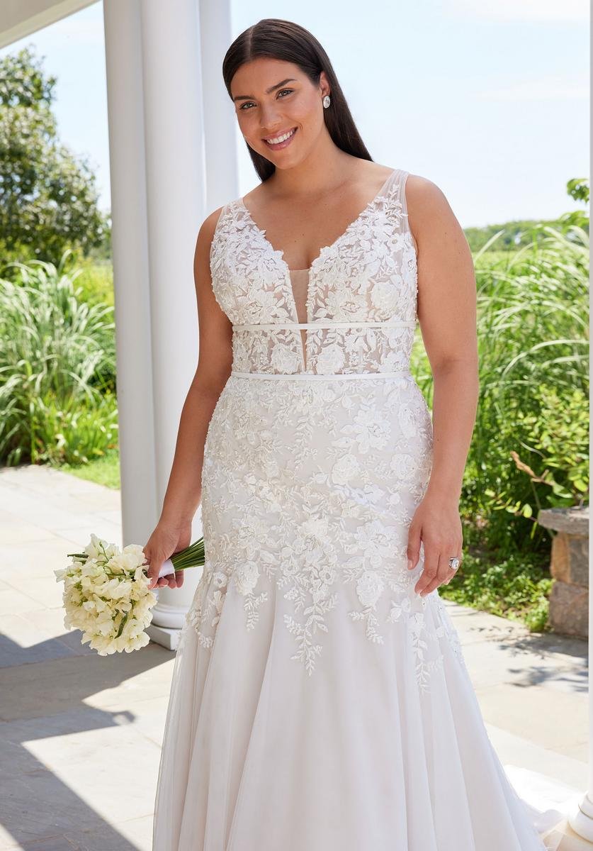 Woman in a white lace wedding dress holding a bouquet of white flowers outdoors, smiling.