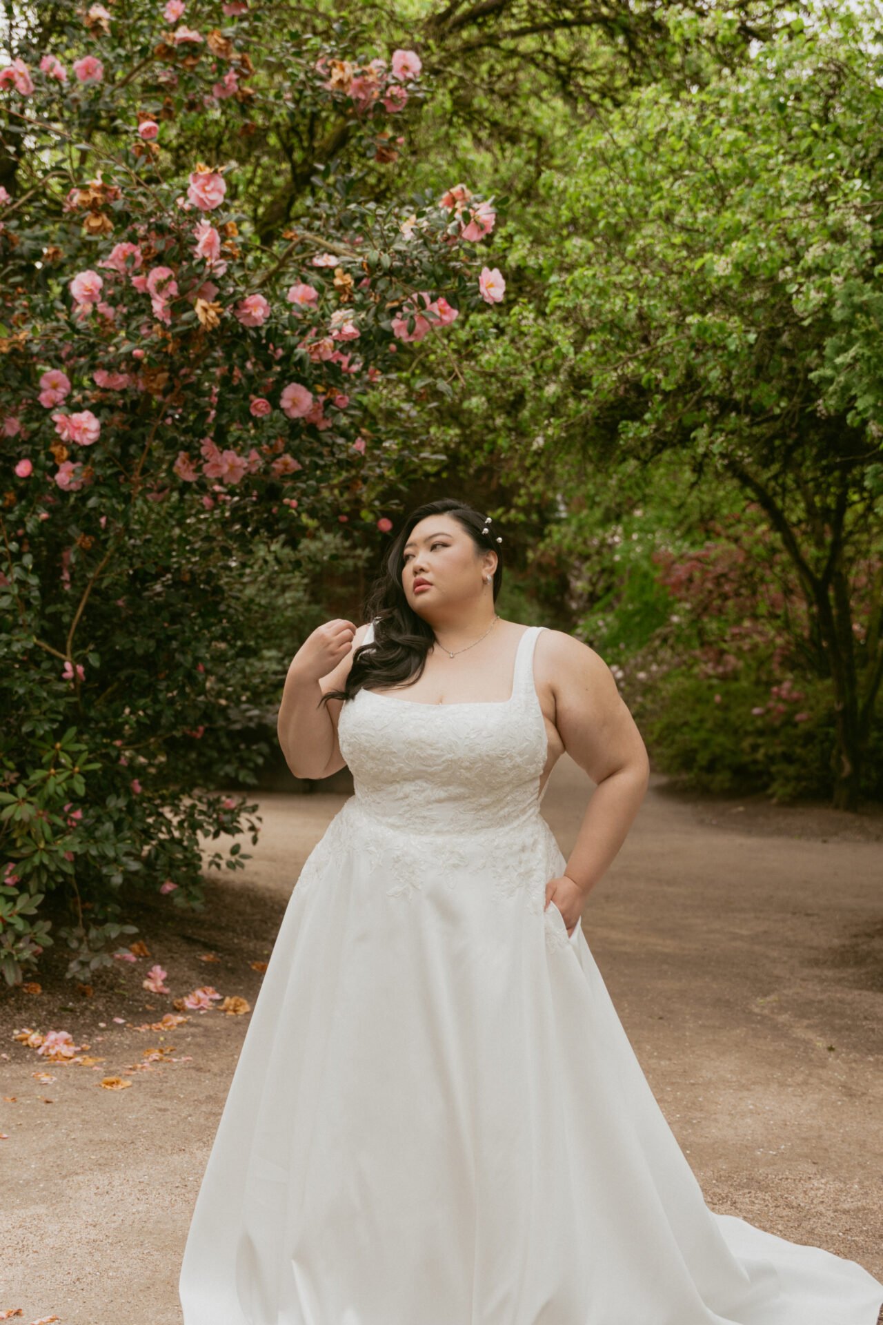 A woman in a white wedding dress standing on a dirt path surrounded by pink and green flowering trees.