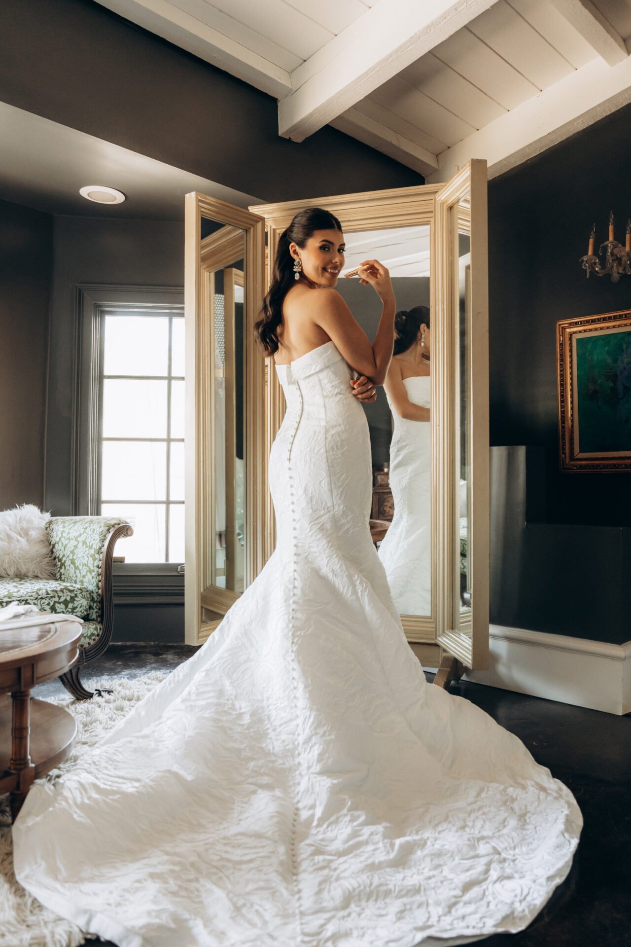 A woman in a strapless white wedding gown standing in front of a mirror, smiling and touching her face, in a room with dark walls and a large window.